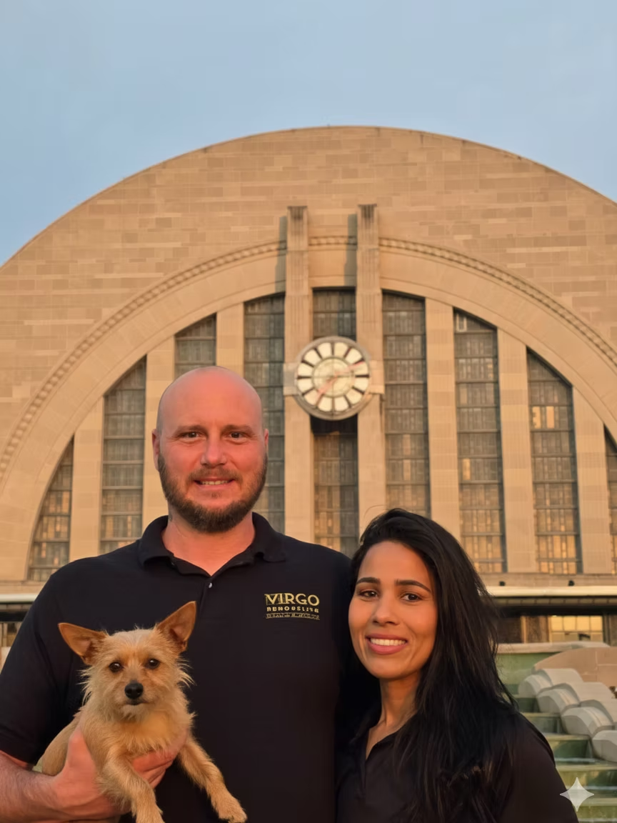 Two people holding a small dog in front of a large arched building at sunset