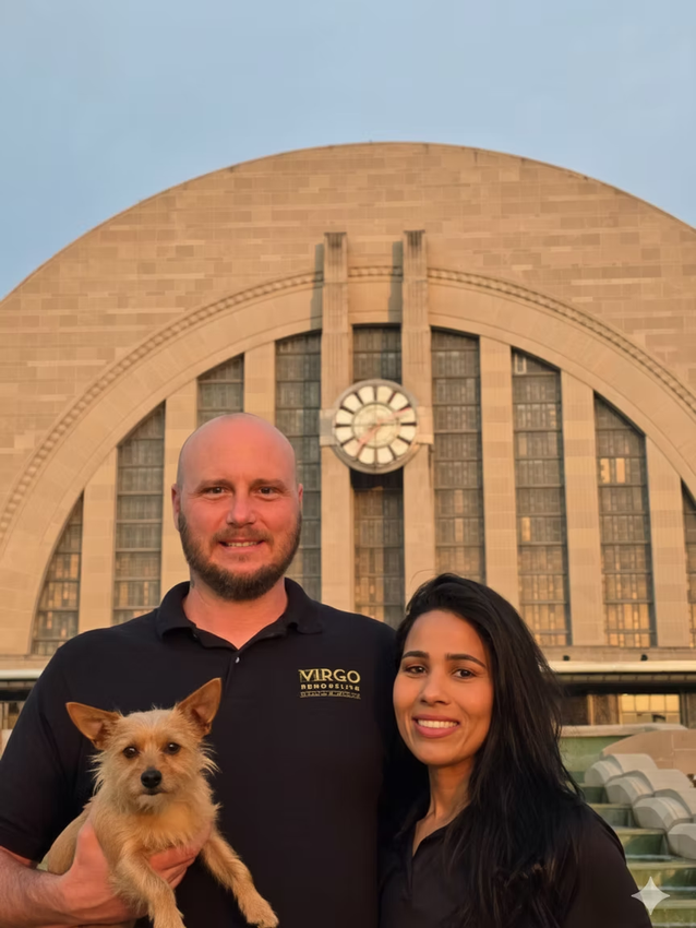 Two people holding a small dog in front of a large arched building at sunset