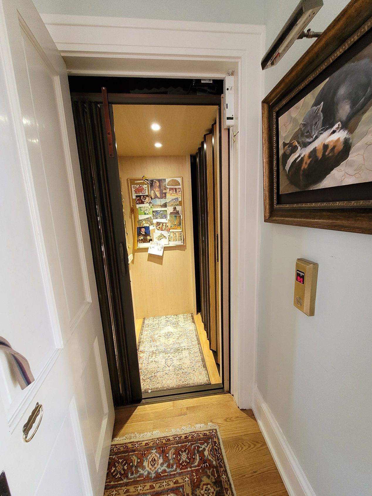 Narrow hallway with rugs, leading to a brightly lit room with a bulletin board and framed painting on the wall