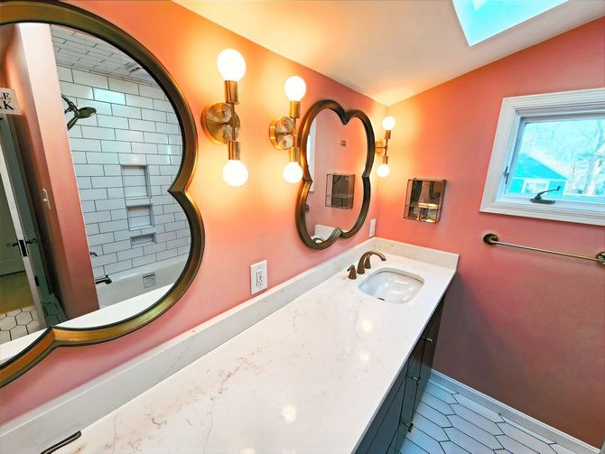 Pink bathroom with a long white sink, round mirrors, wall sconces, and skylight lighting.