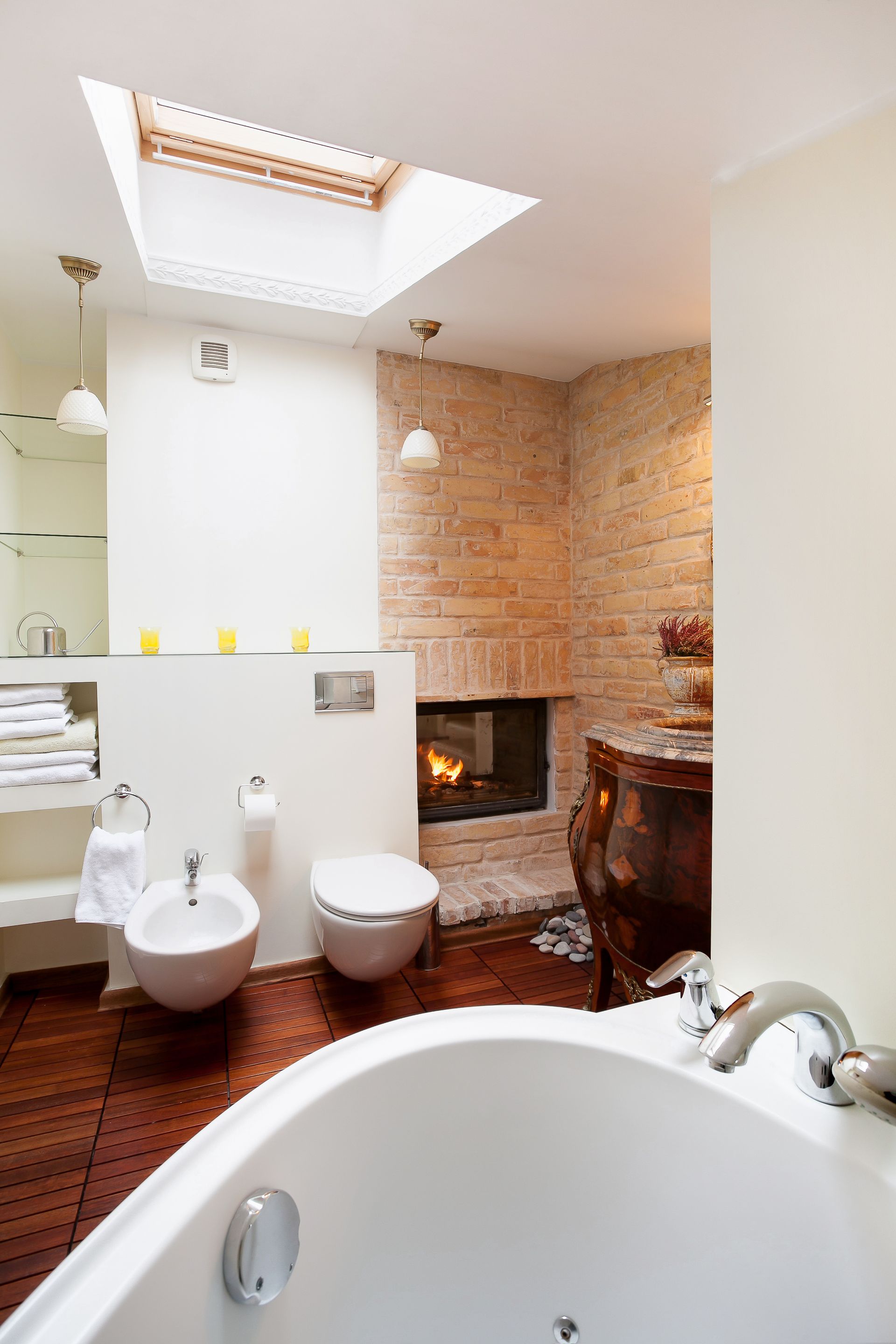 Bright bathroom with a skylight, freestanding tub, toilet, and a fireplace by the brick wall