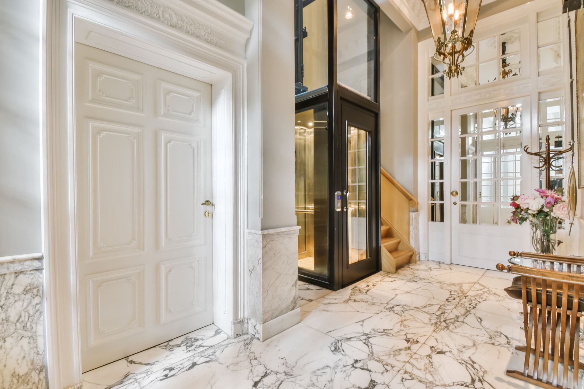 Elegant marble foyer with white paneled doors, glass elevator, and a chandelier, with a small table and flowers