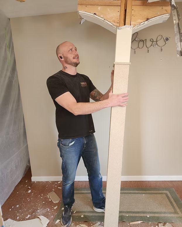 Man holding a porch column in a room with debris on the floor and exposed ceiling beams
