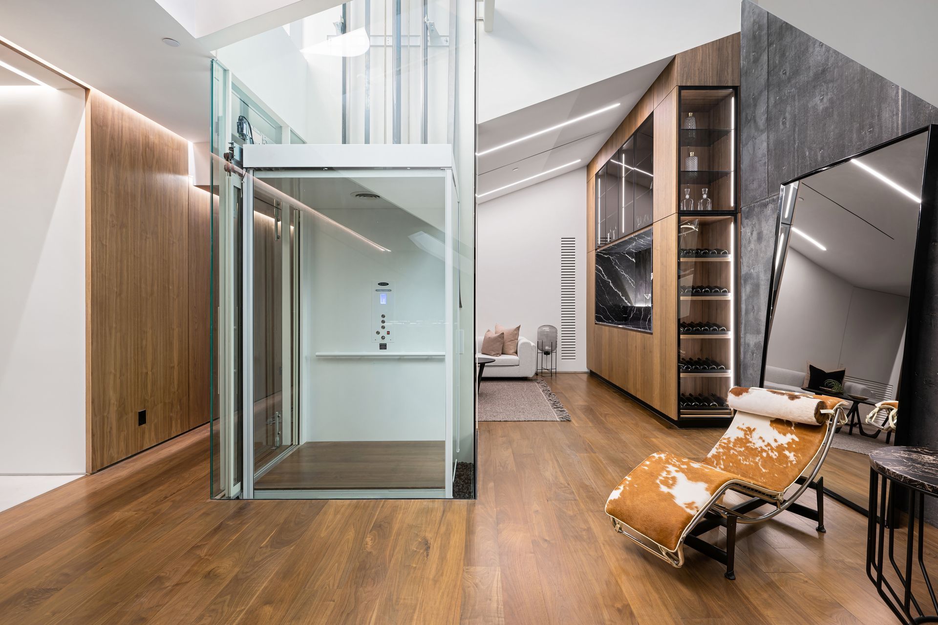 Modern open-plan interior with wood floors, glass elevator, and lounge seating under a staircase