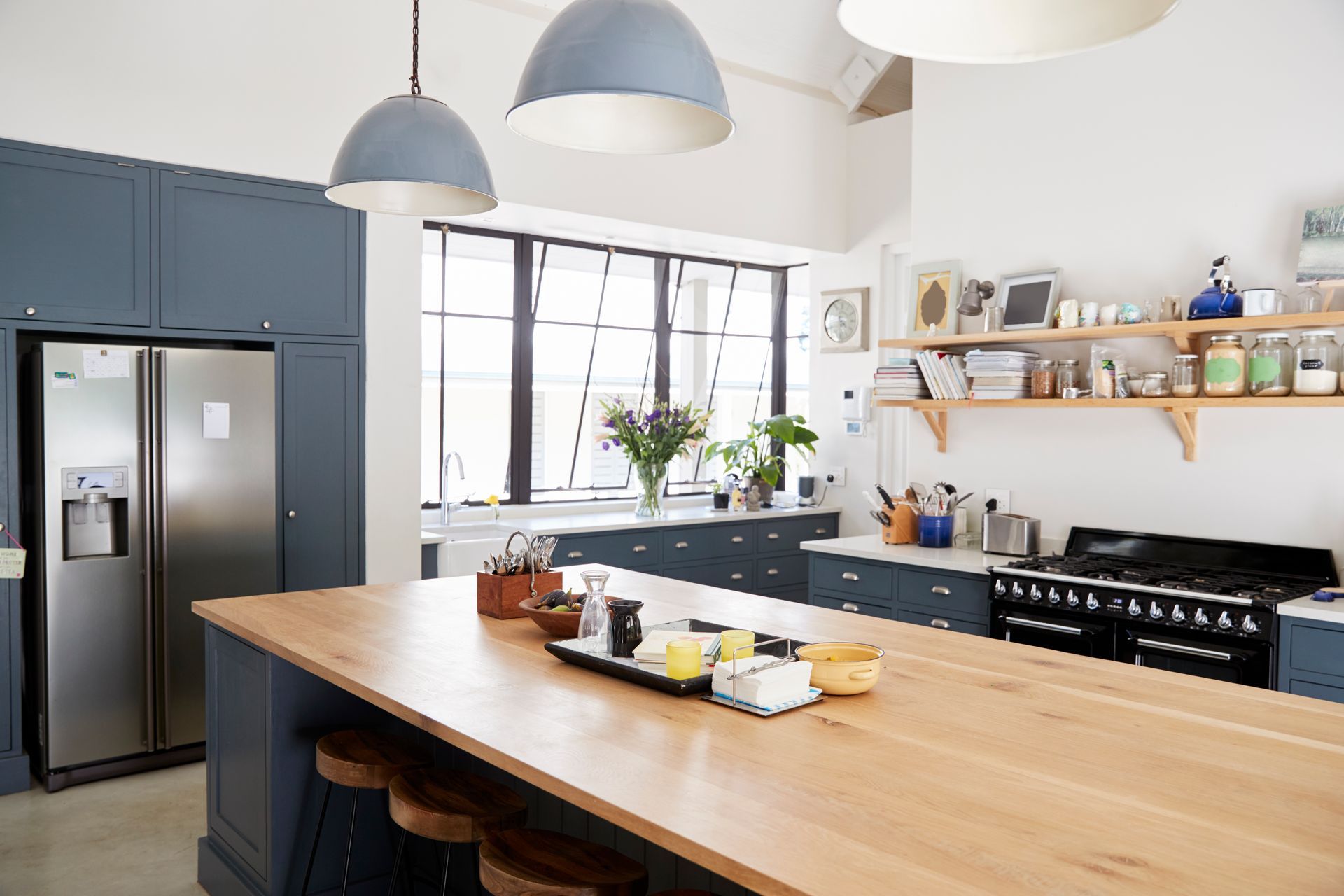 Modern kitchen with wooden island, stainless fridge, pendant lights, and open shelves by large windows