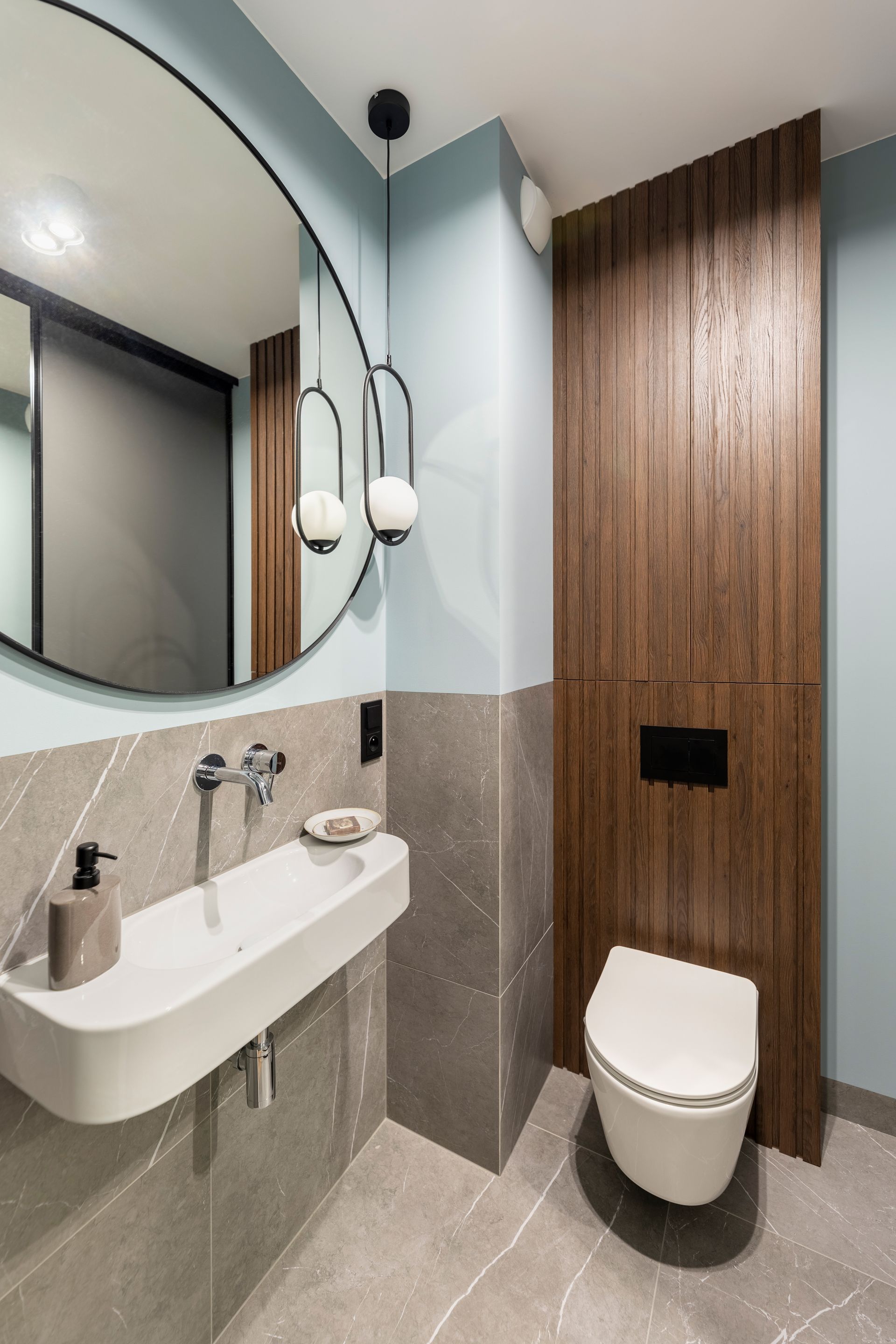 Modern compact bathroom with round mirror, wall sink, toilet, and wood-paneled partition
