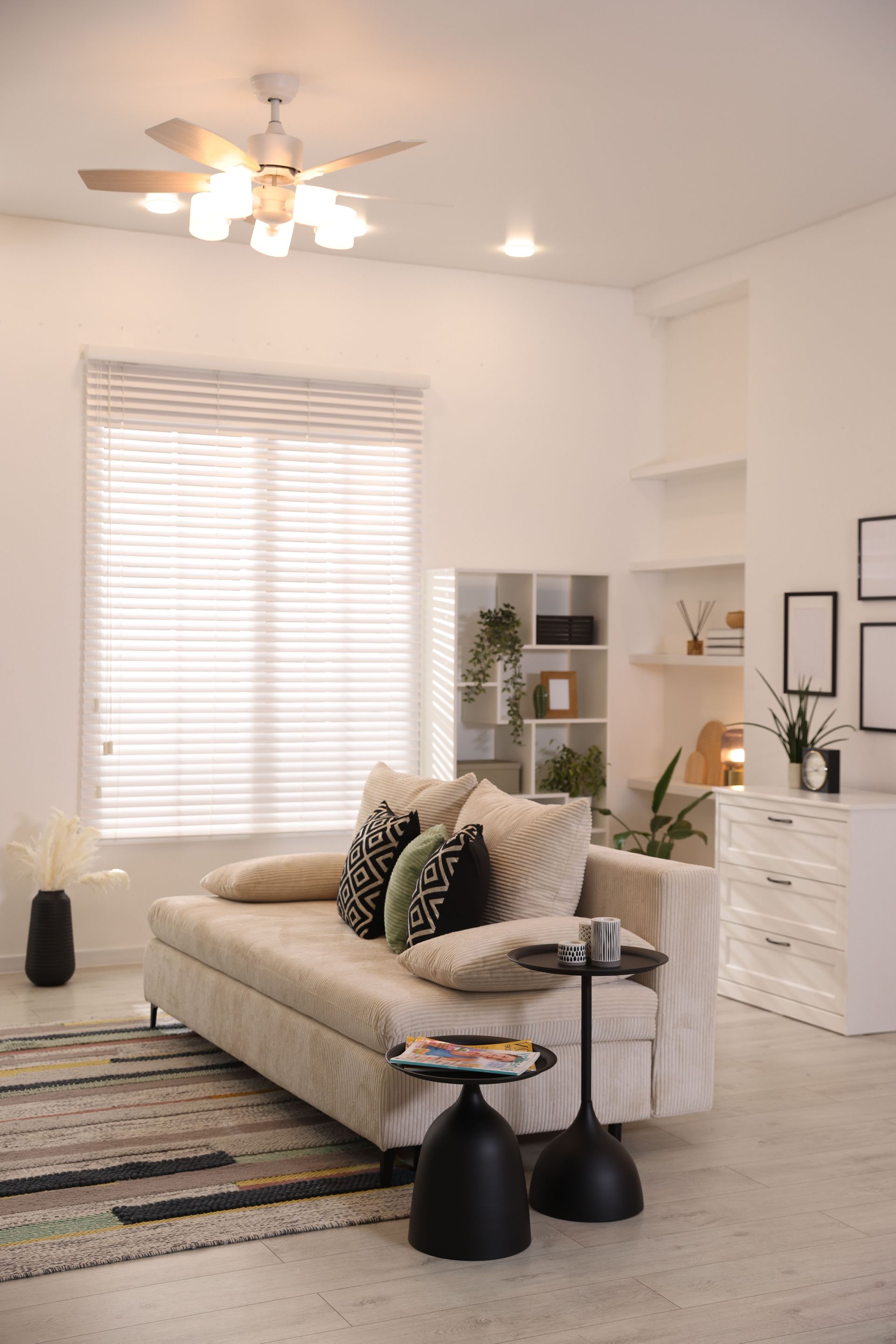 Bright living room with beige sofa, black accent tables, striped rug, white dresser, and large window blinds.