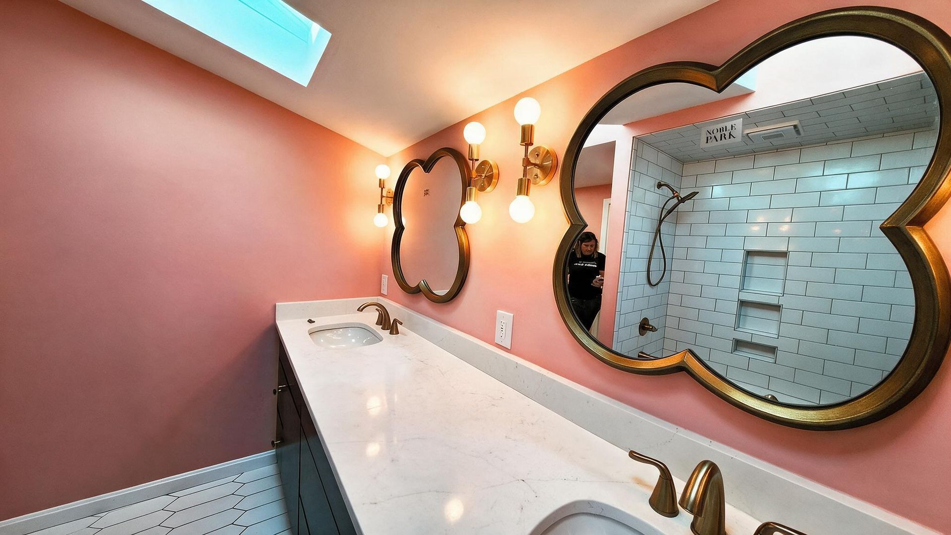 Elegant bathroom with pink walls, long white vanity, gold-framed mirror, and wall sconces.
