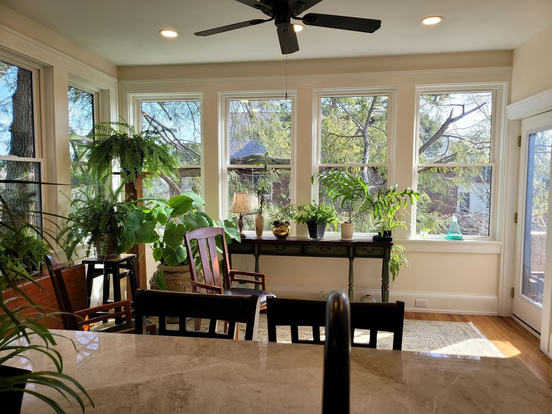 Bright sunroom with large windows, indoor plants, wooden chairs, and a dining table.