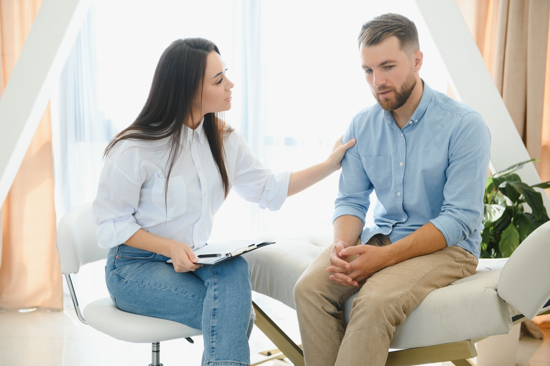 A therapist comforts a man in a light-filled room. The therapist, wearing a white shirt and jeans,