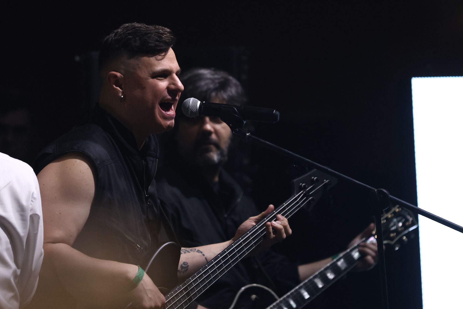 Dois músicos se apresentando ao vivo no palco, um cantando ao microfone enquanto toca baixo, o outro tocando guitarra.