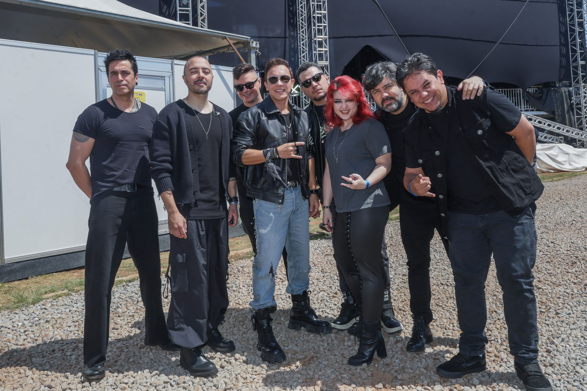 Sete pessoas posando juntas ao ar livre, vestidas principalmente de preto, em frente a um palco.