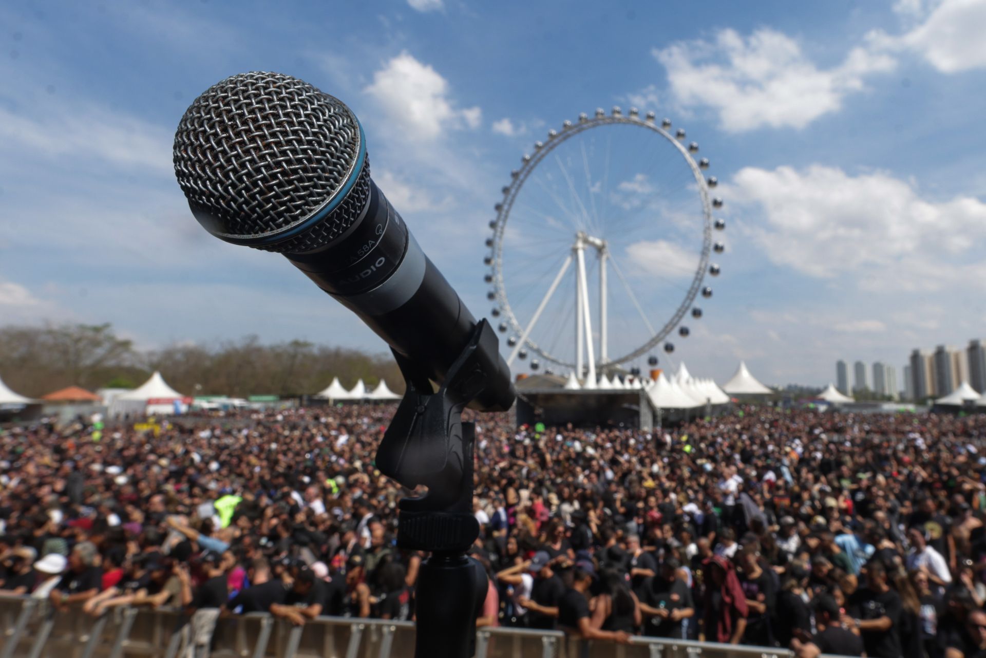 Microfone em primeiro plano, voltado para uma grande multidão em um show ao ar livre, com uma roda-gigante ao fundo.
