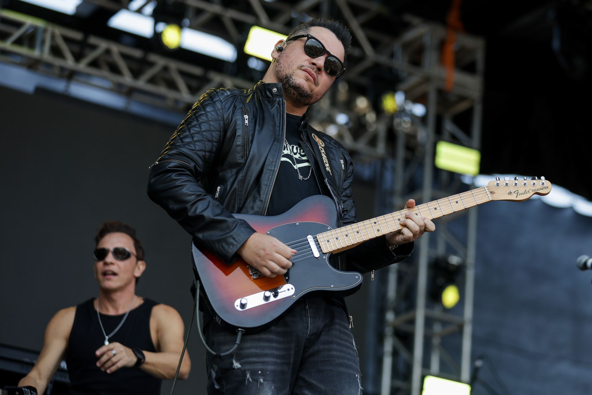 Um guitarrista de jaqueta de couro toca uma guitarra elétrica sunburst no palco, com um músico de regata ao lado.