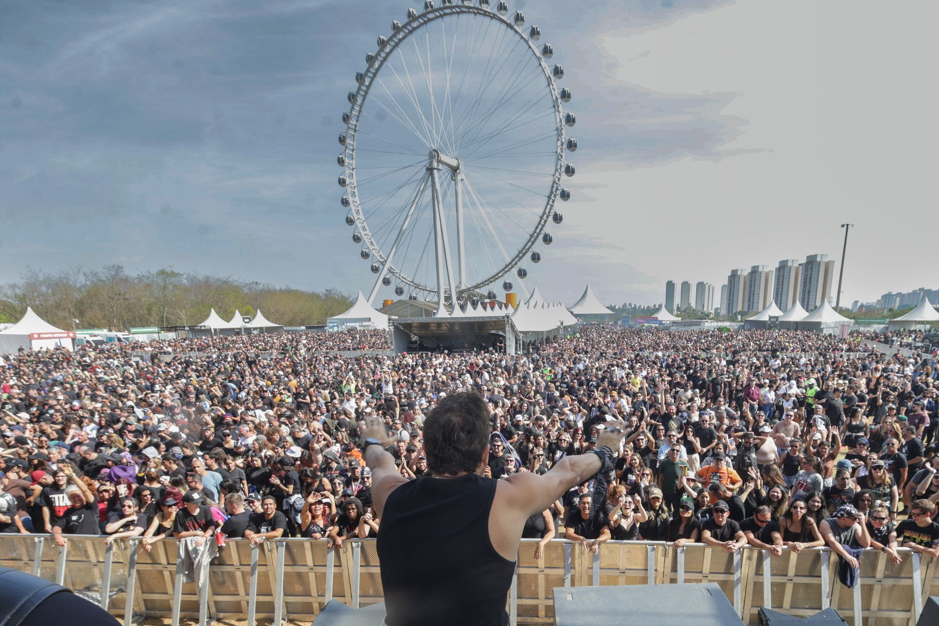 Público em um show ao ar livre, visto do palco com uma roda-gigante ao fundo.