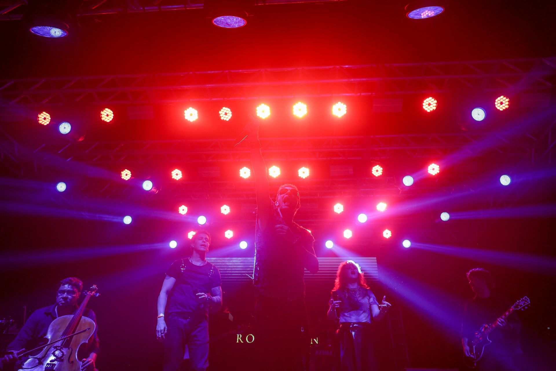Três pessoas no palco, cantando. Luzes de palco roxas e vermelhas as iluminam.
