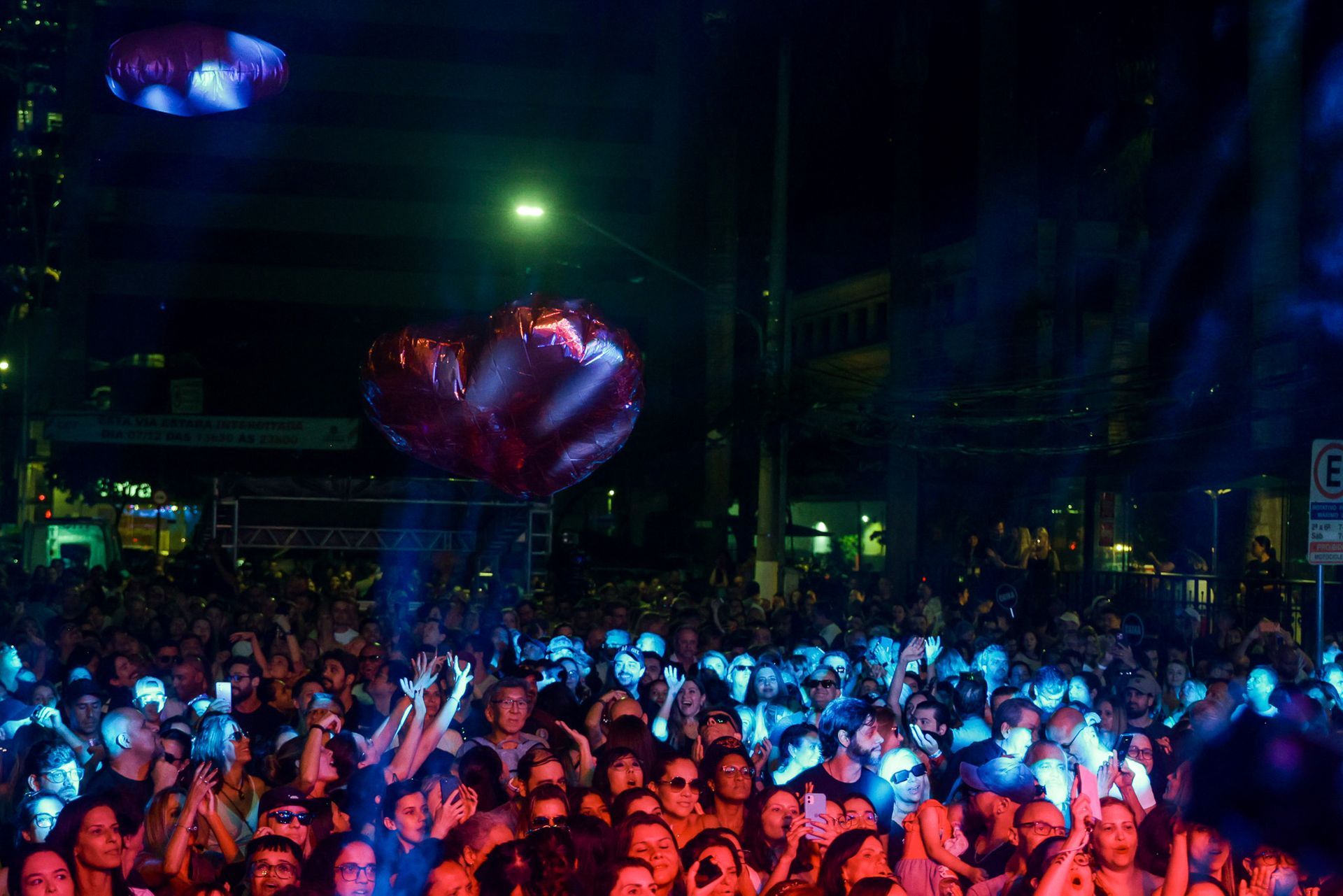 Grande público em show ao ar livre, iluminado por luzes de palco azuis e vermelhas, com balões em forma de coração