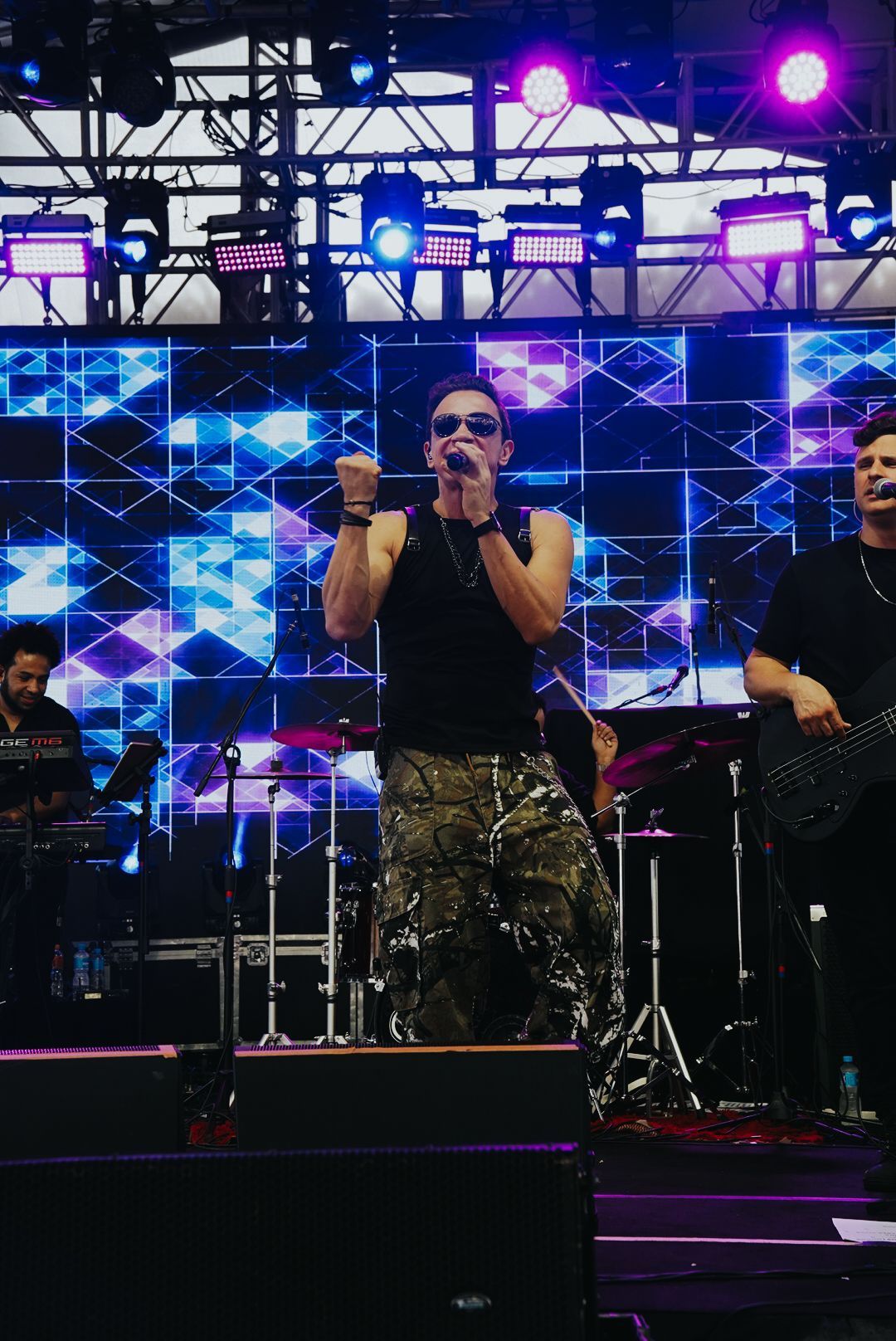 Banda se apresentando no palco, vocalista com o punho erguido, calça camuflada, fundo iluminado.