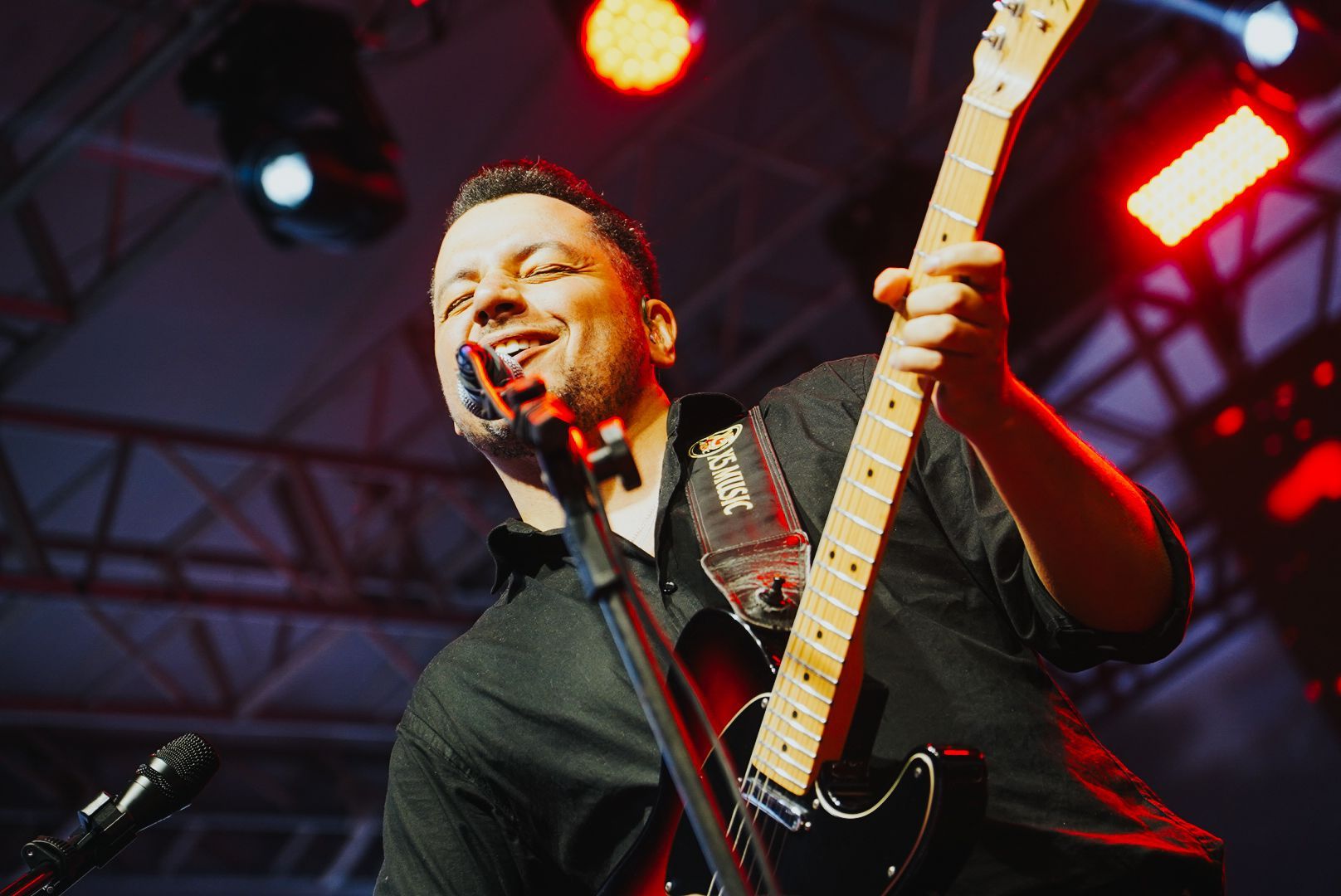 Um artista tocando guitarra elétrica e cantando ao microfone no palco, sob luzes de palco.