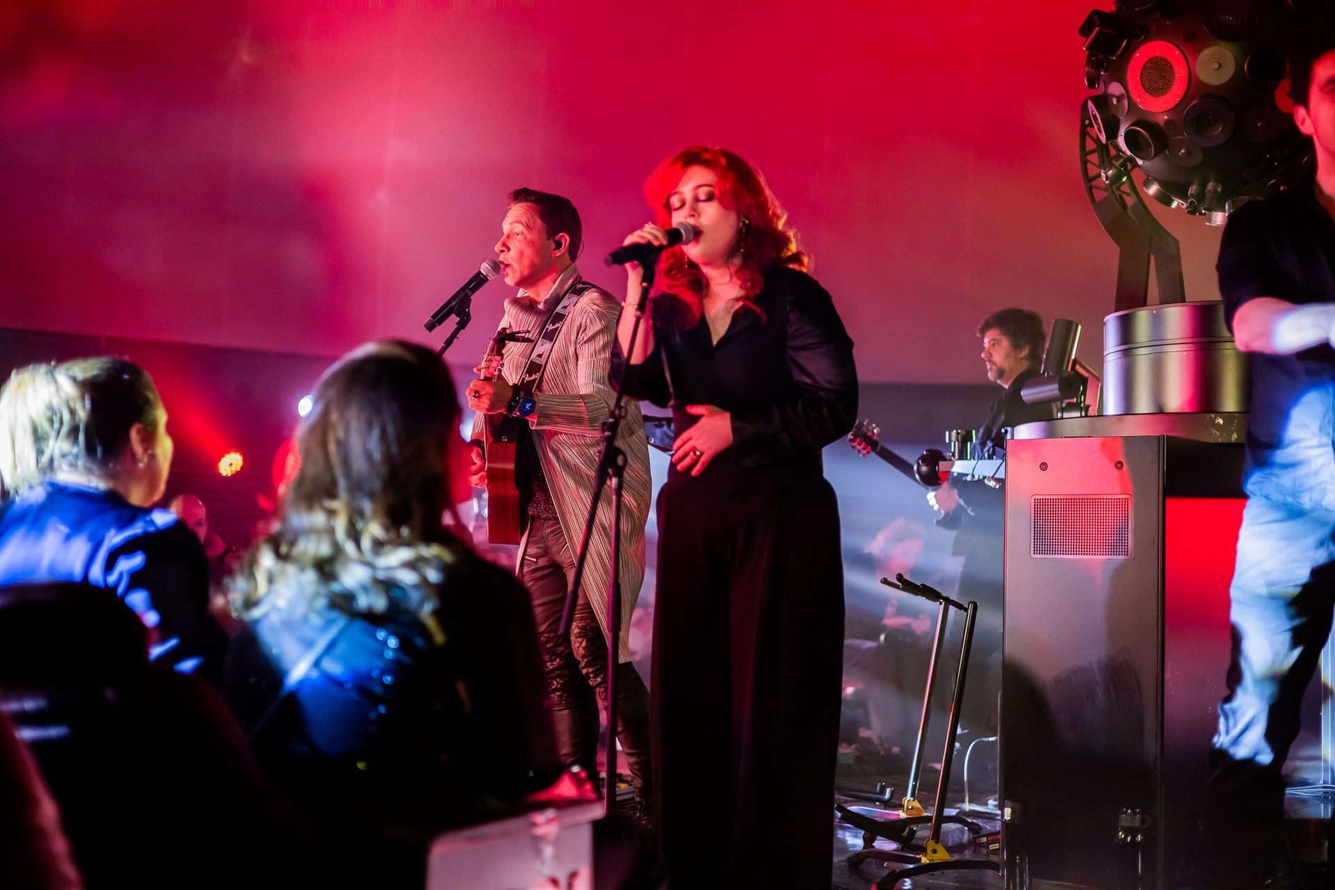 Um grupo musical se apresenta em um palco banhado por luz vermelha, com uma mulher cantando e um guitarrista ao seu lado.