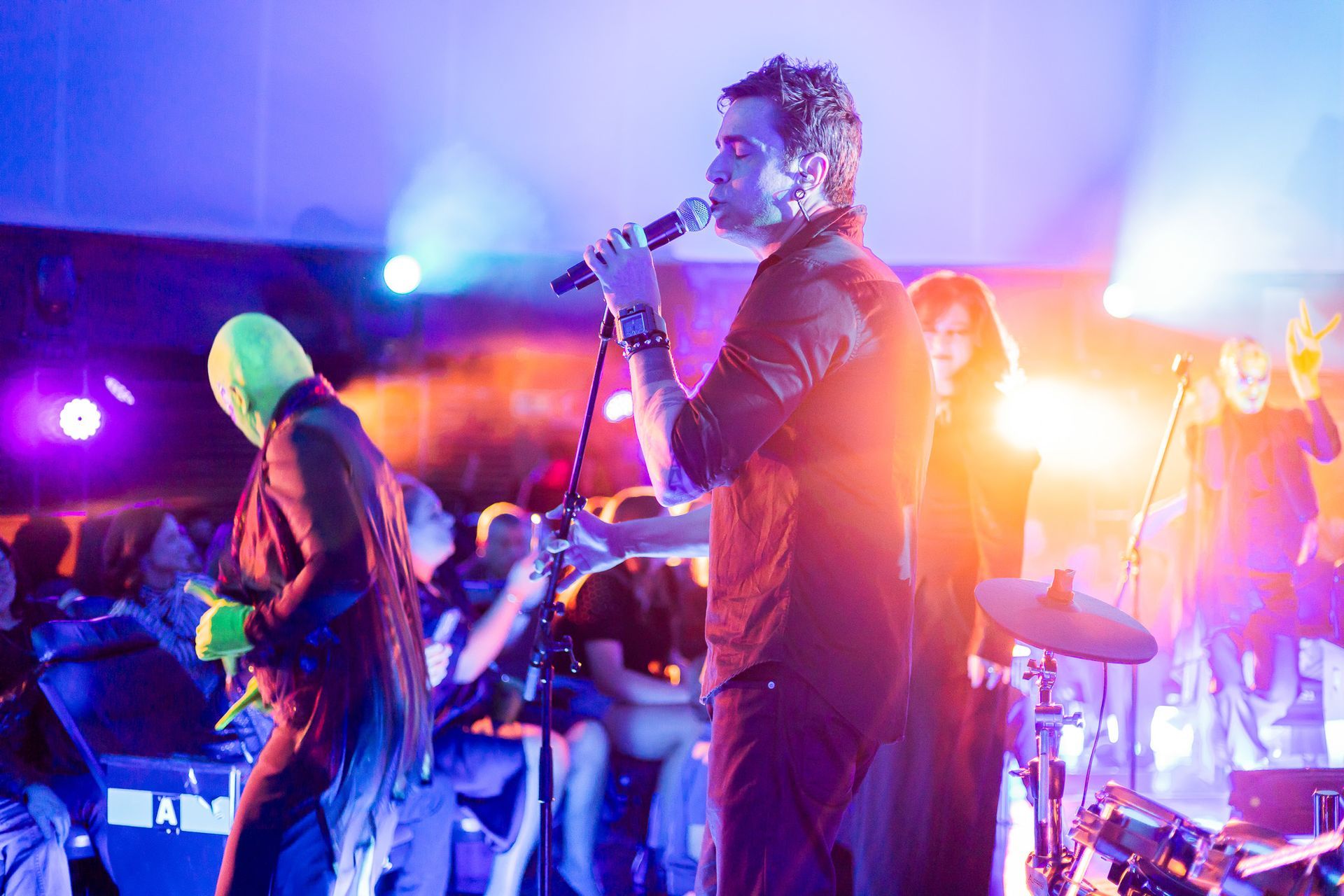 Uma banda se apresenta no palco sob luzes brilhantes e coloridas, com um cantor e um músico usando uma máscara verde.