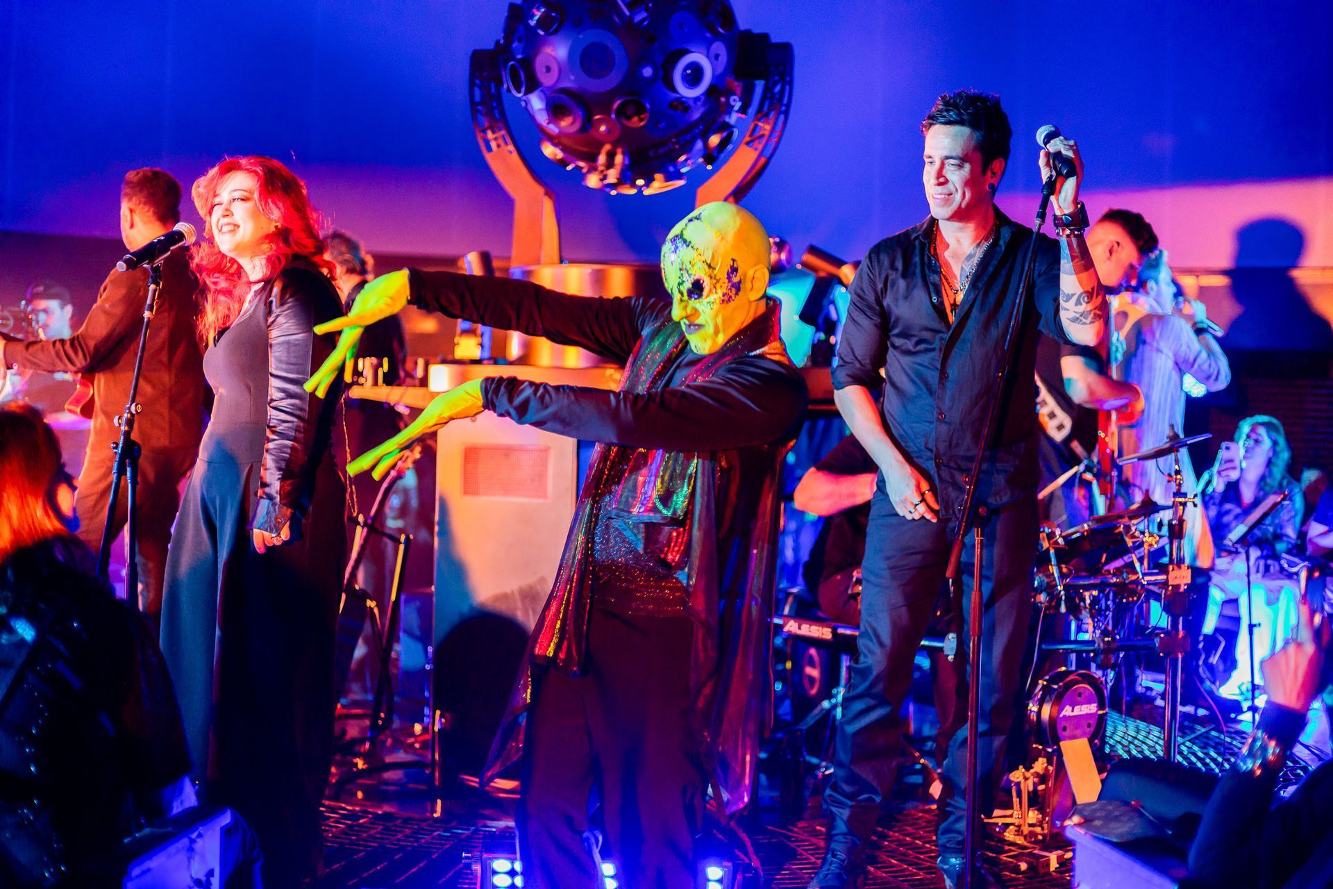 Uma banda se apresenta em um palco banhado por luz azul, com um dos artistas usando máscara e luvas verde neon.