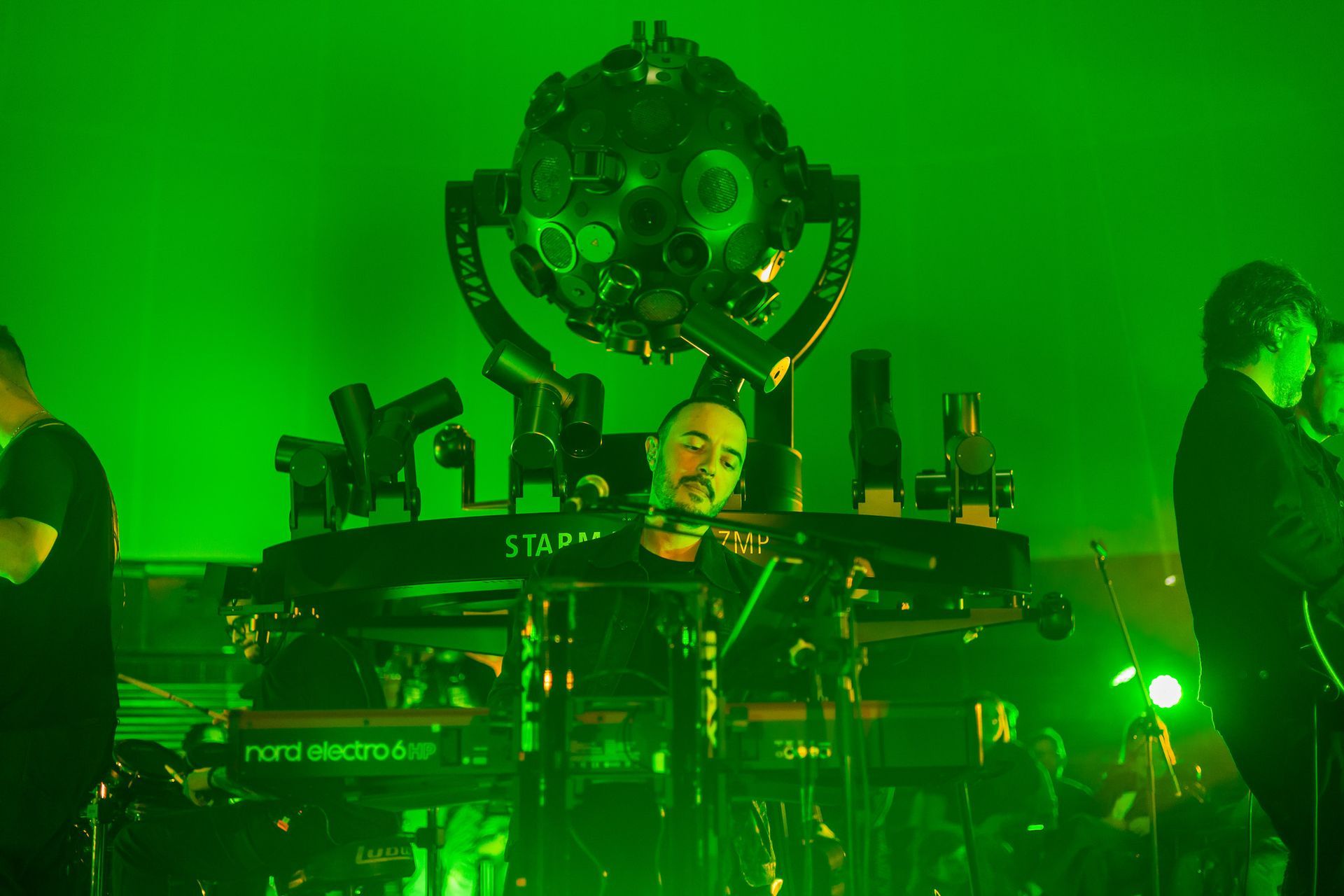 Um músico toca teclado no palco sob uma iluminação verde brilhante, abaixo de uma grande estrutura de luz esférica.