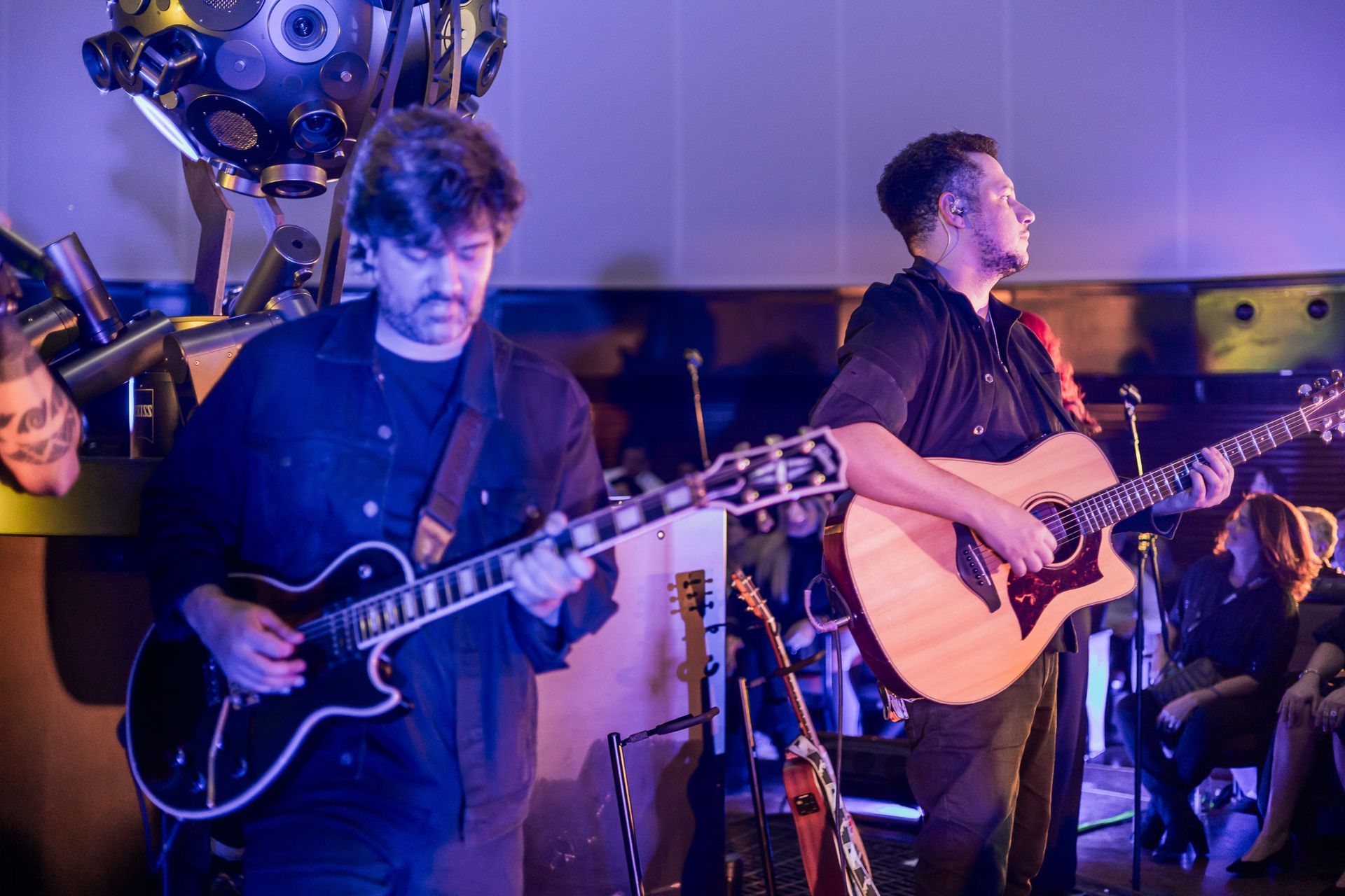 Dois músicos se apresentam no palco; um toca uma guitarra elétrica com som grave, enquanto o outro toca um violão.