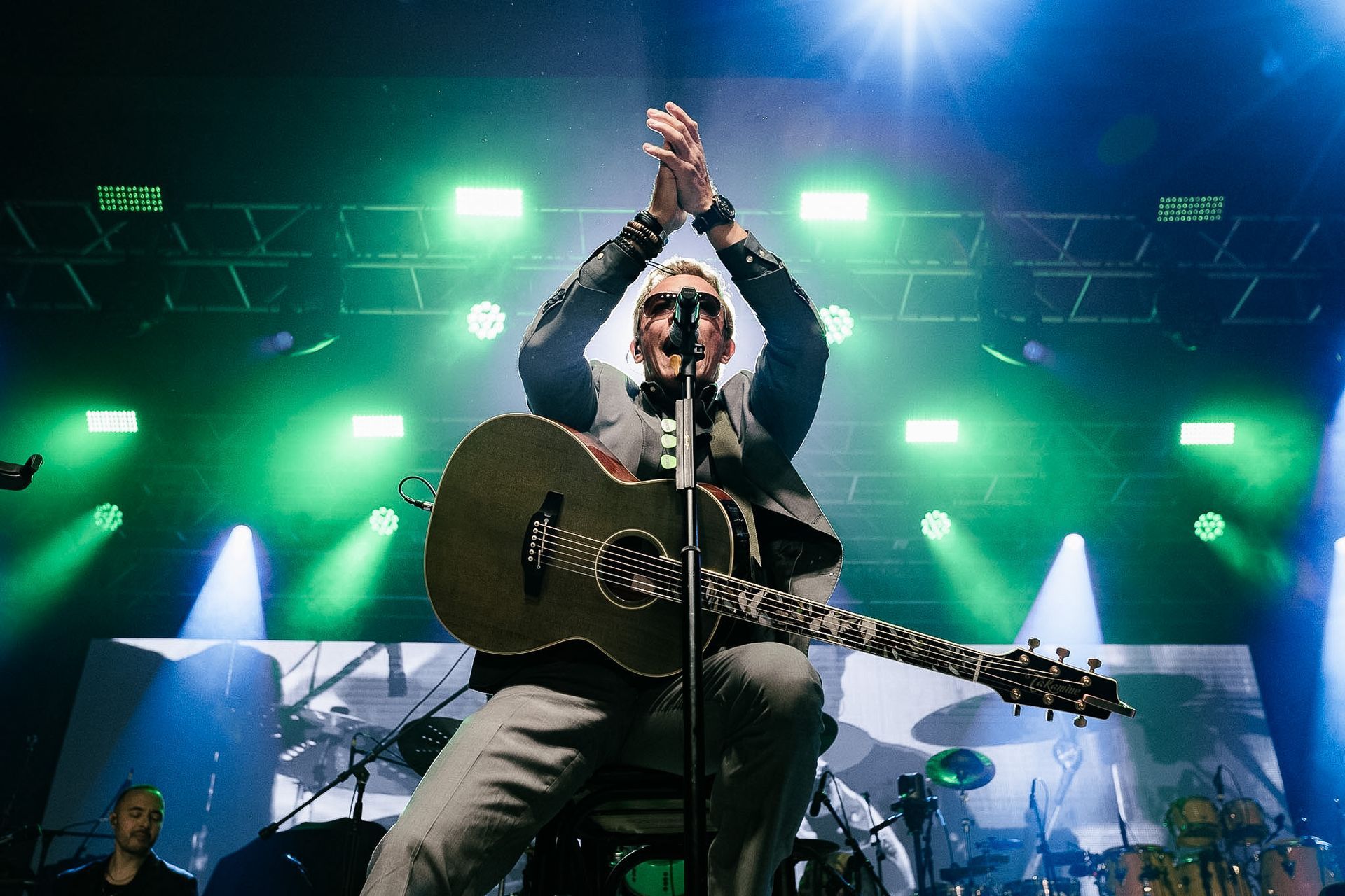 Músico com guitarra no palco sob luzes verdes, batendo palmas acima da cabeça durante um show ao vivo.