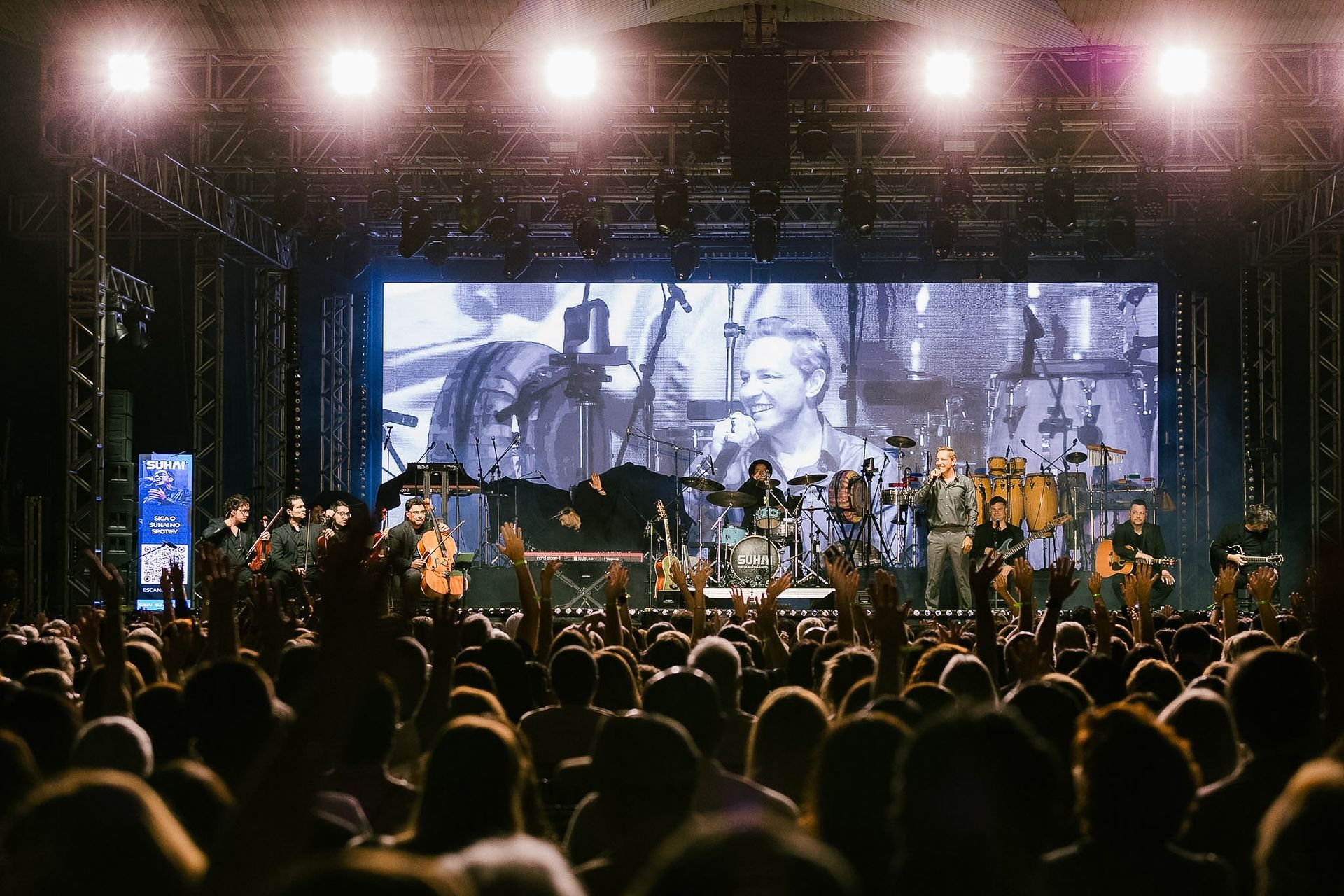Palco de concerto ao ar livre com luzes brilhantes, banda tocando e público assistindo em primeiro plano.