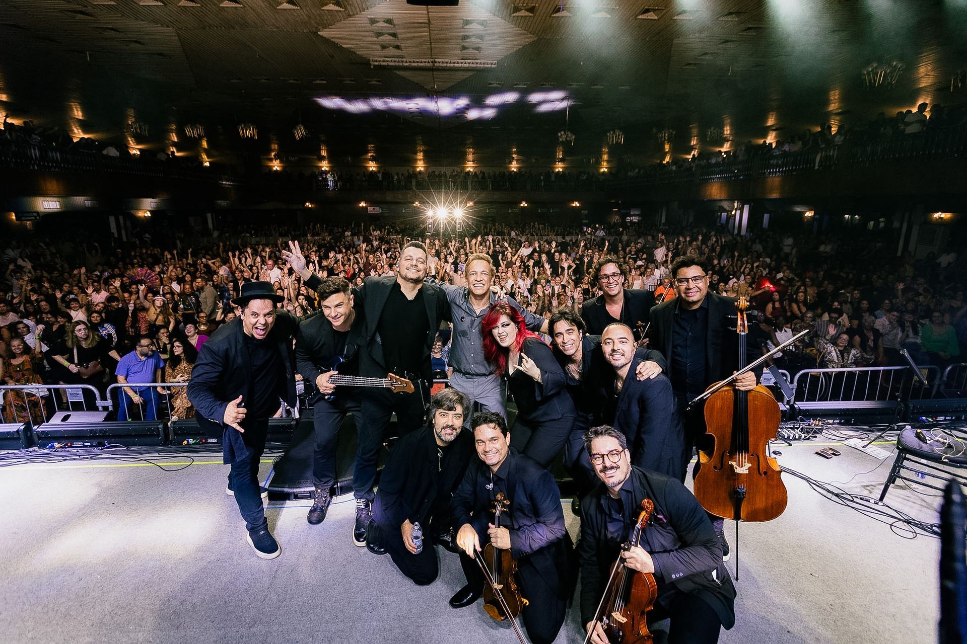 Grupo de músicos posando no palco diante de uma grande plateia entusiasmada sob luzes quentes de palco.