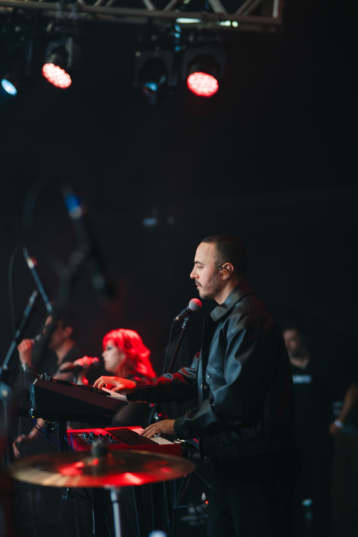 Um homem toca teclado e canta no palco, iluminado por luzes vermelhas, com os outros membros da banda visíveis.