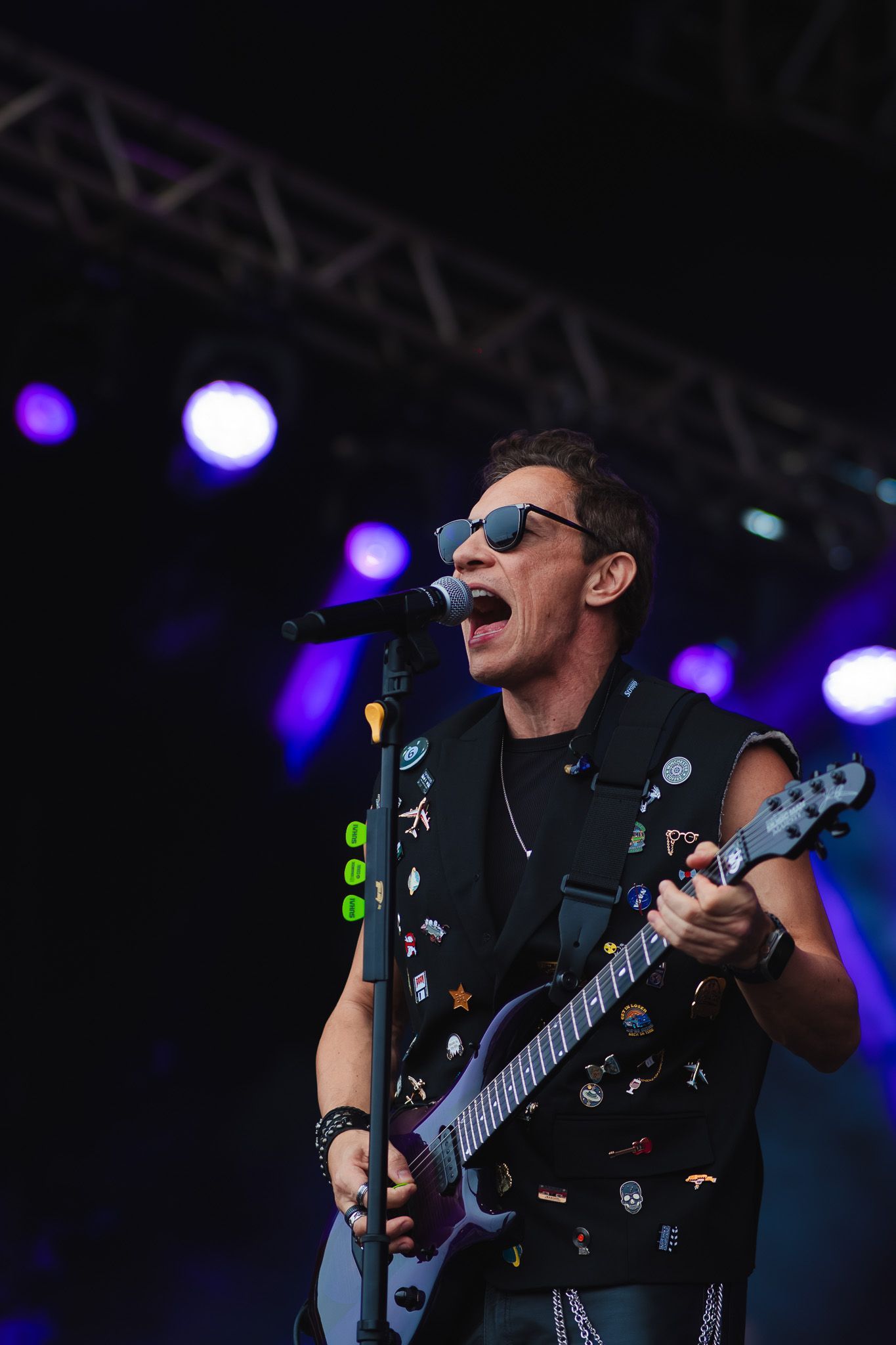 Um artista cantando e tocando guitarra elétrica no palco, usando óculos escuros e um colete com tachas, sob luzes de palco.
