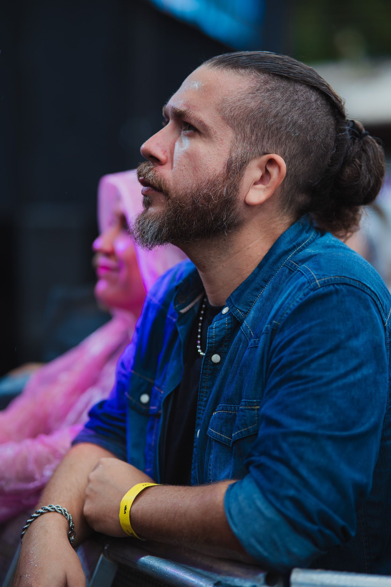 Homem com as laterais raspadas, barba e coque, olhando para cima, vestindo uma camisa jeans azul, assistindo a um evento.