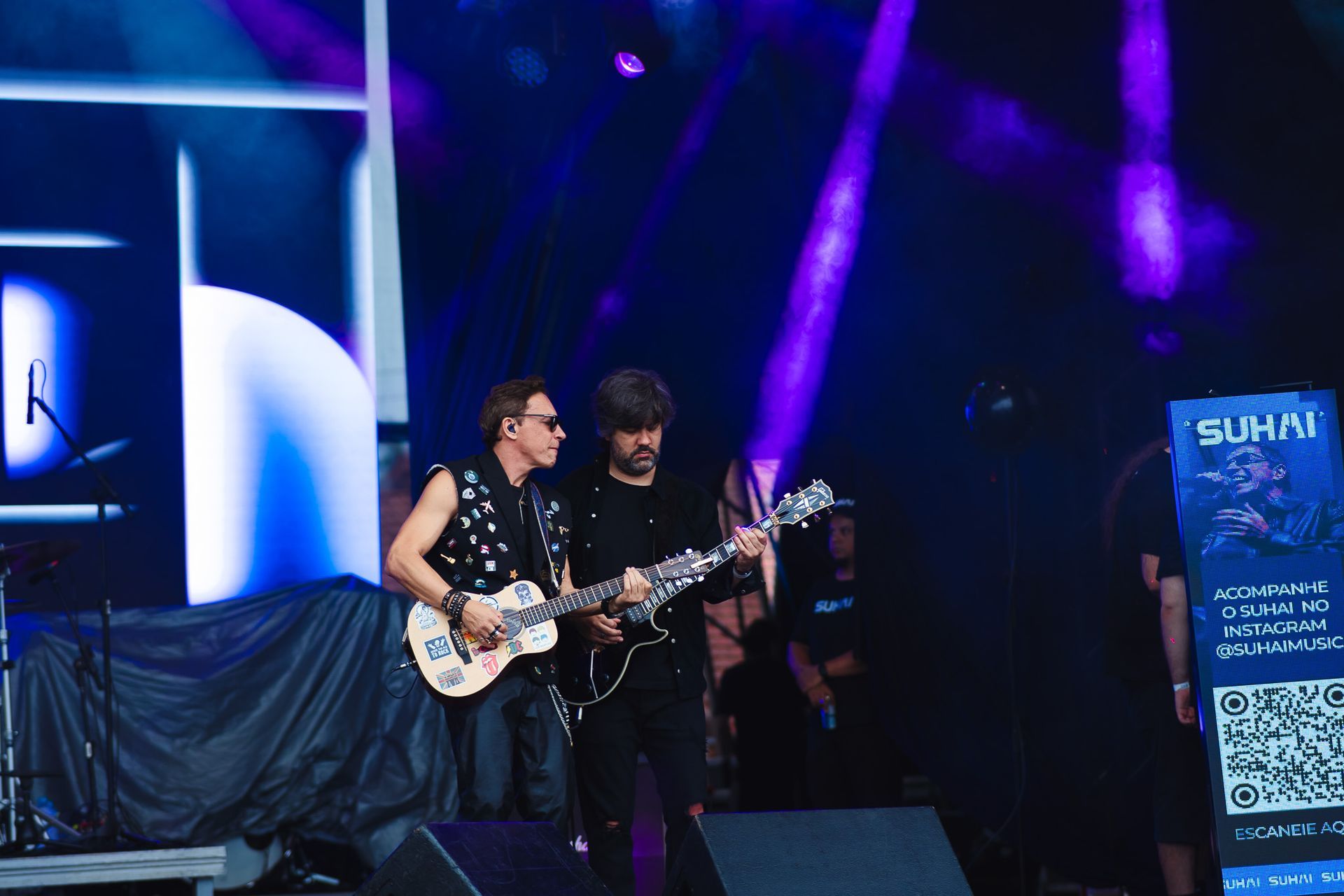 Dois músicos tocando guitarras elétricas no palco sob luzes roxas.