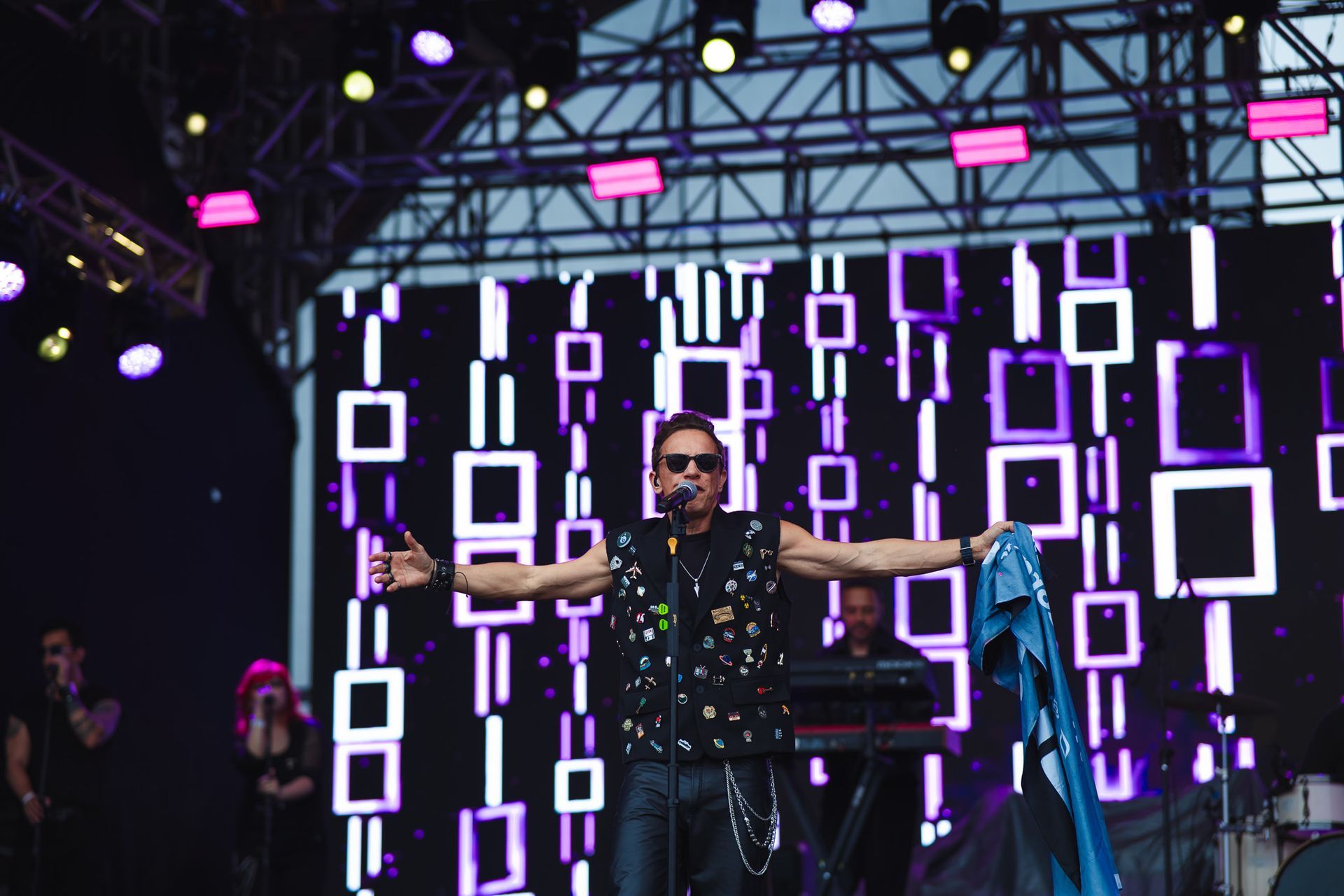 Um artista no palco com os braços estendidos, usando óculos escuros e colete preto.