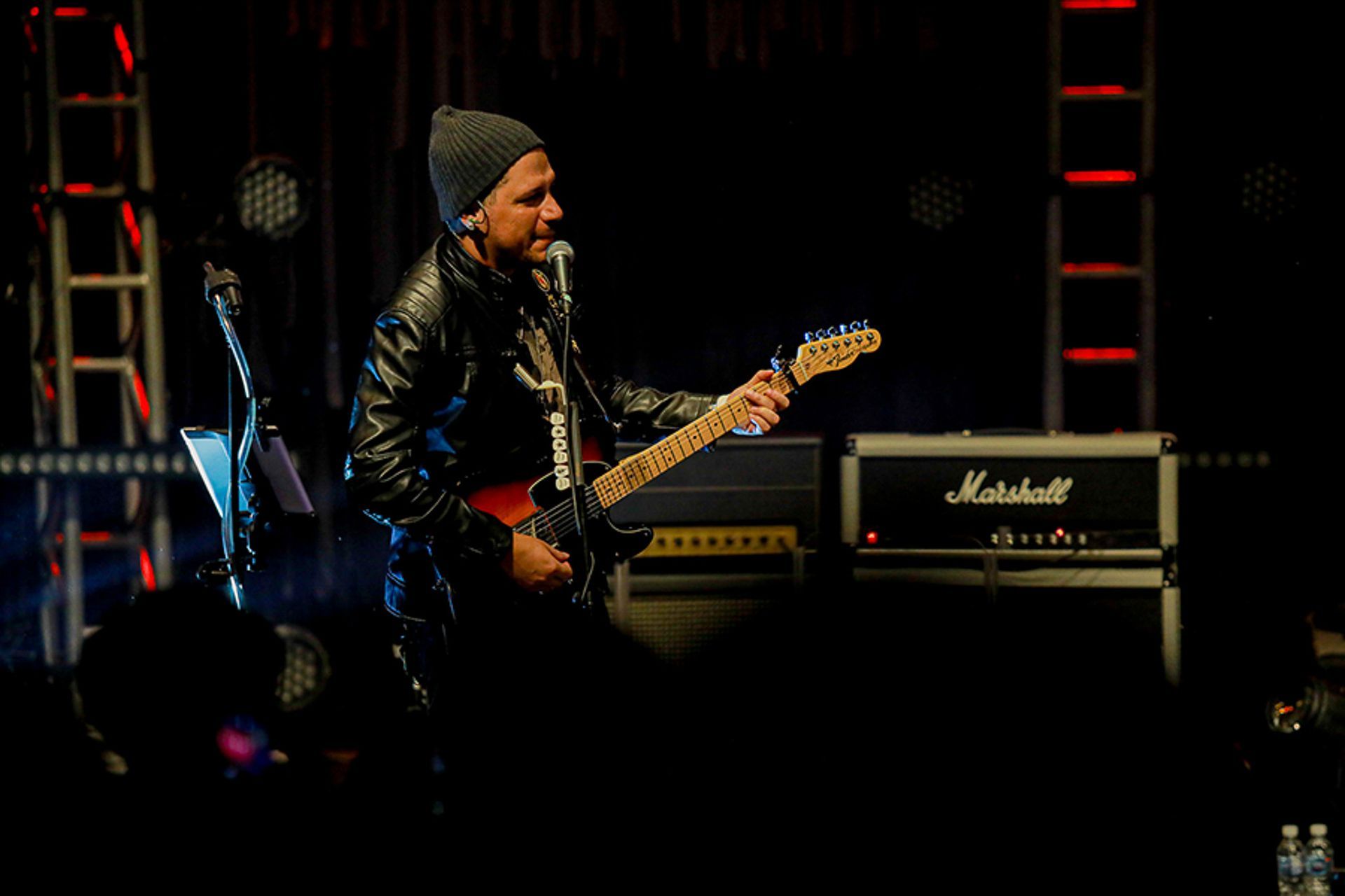 Um artista de gorro e jaqueta de couro toca guitarra elétrica enquanto canta ao microfone em um palco de shows.