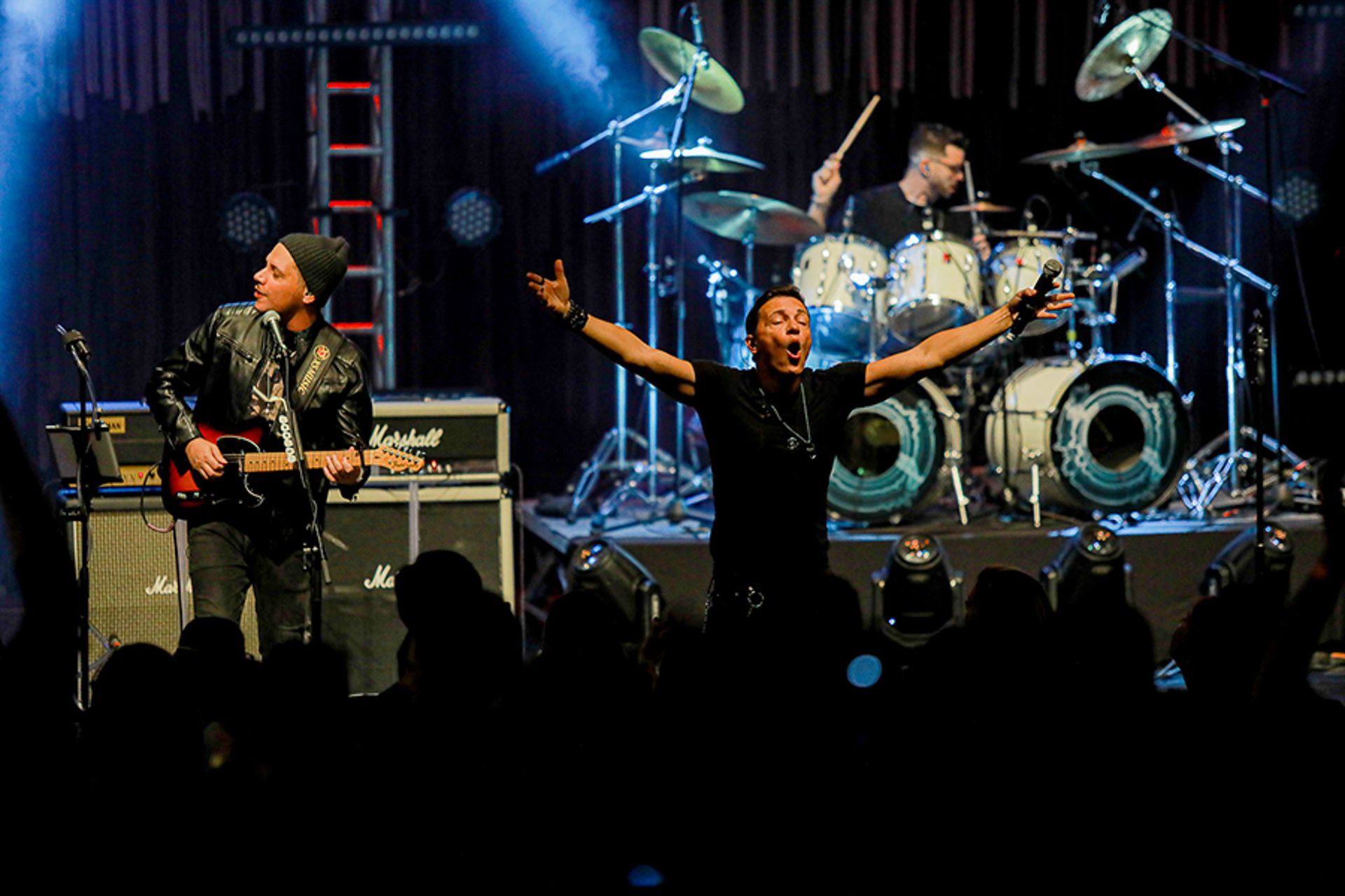 Uma banda de rock se apresenta no palco: um guitarrista toca, o vocalista permanece de braços abertos e um baterista.