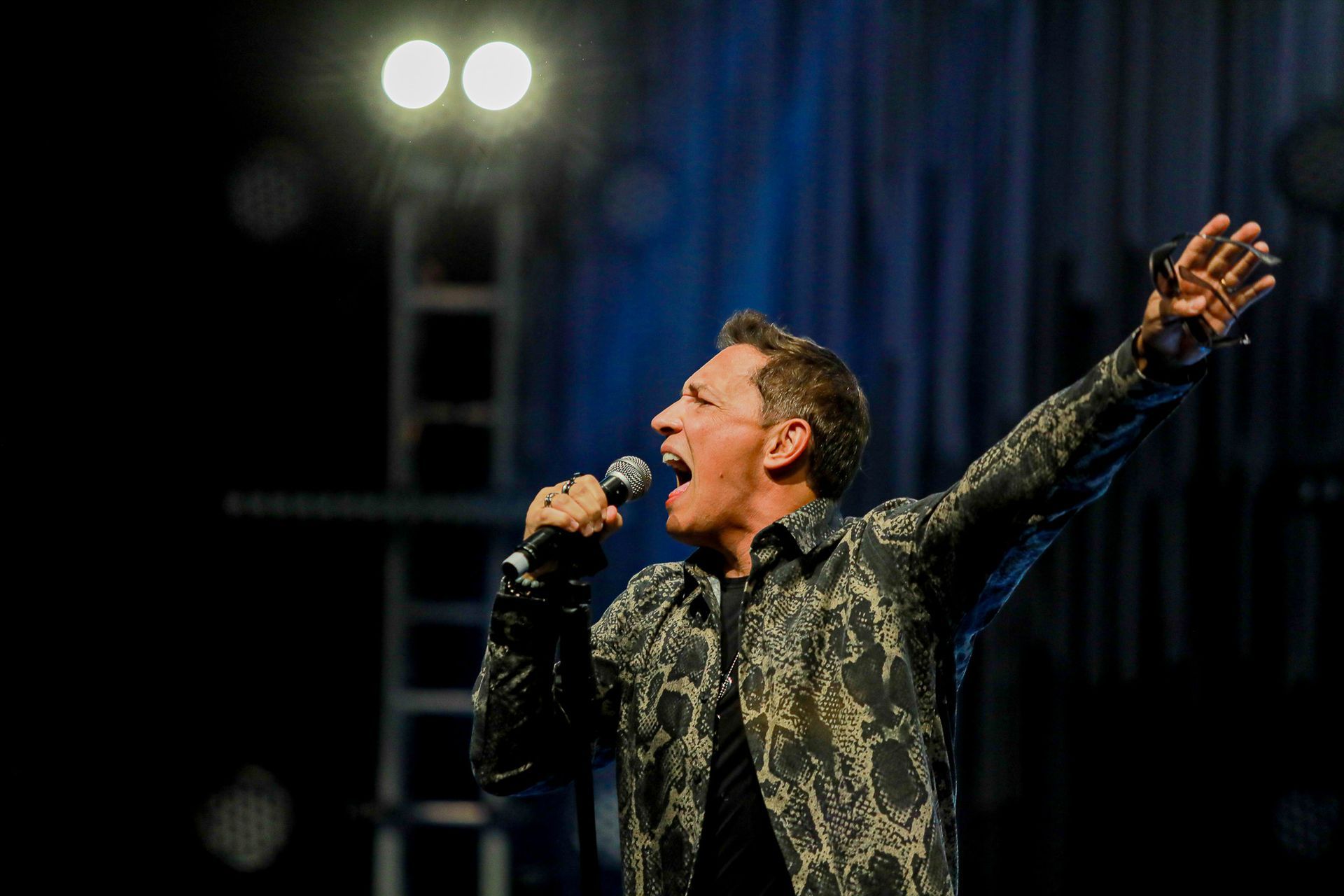 Um cantor cantando em um microfone no palco, vestindo uma camisa escura estampada, com um braço erguido em direção à plateia.