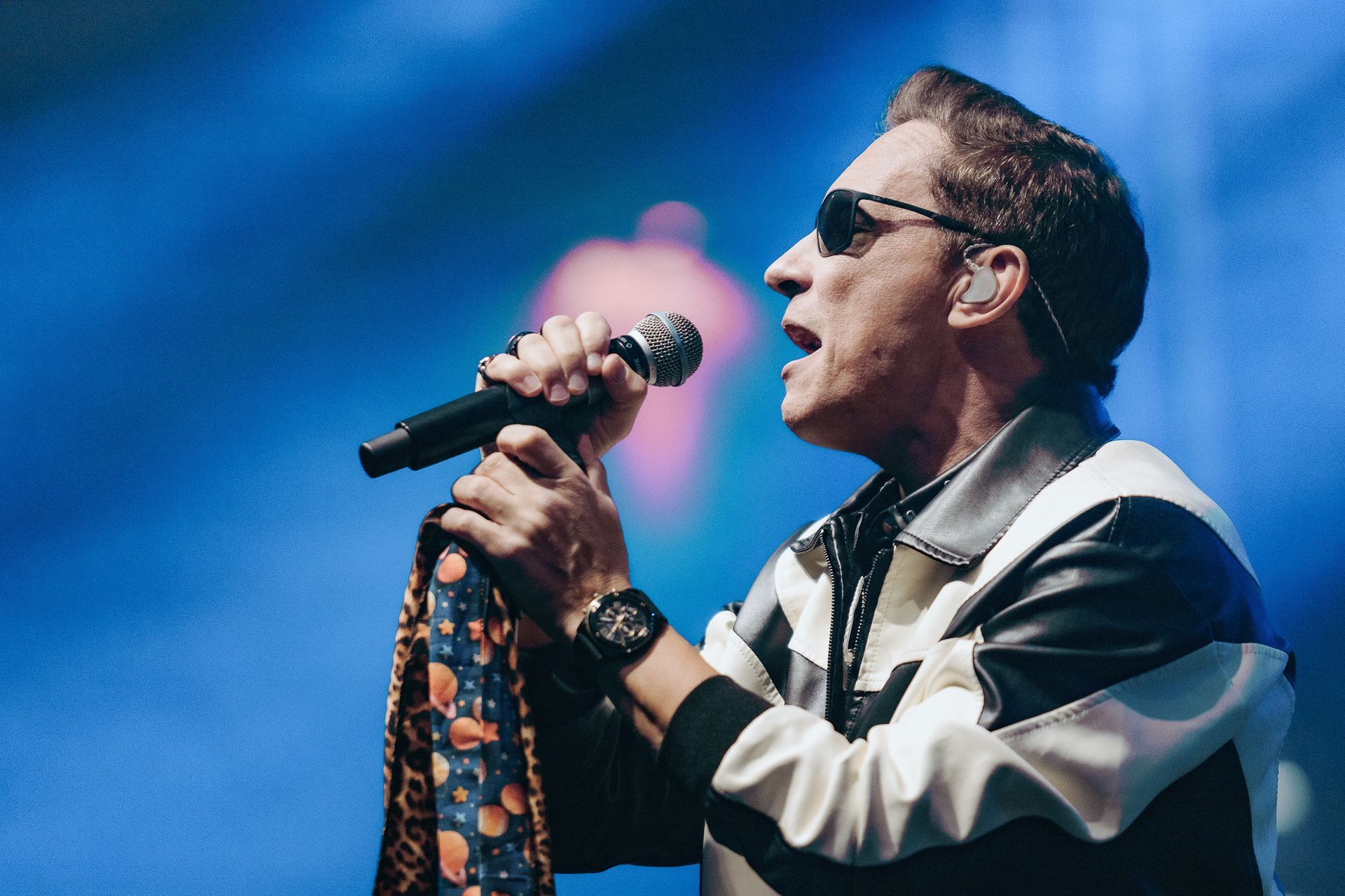 Cantor de óculos escuros e lenço estampado cantando ao microfone no palco.