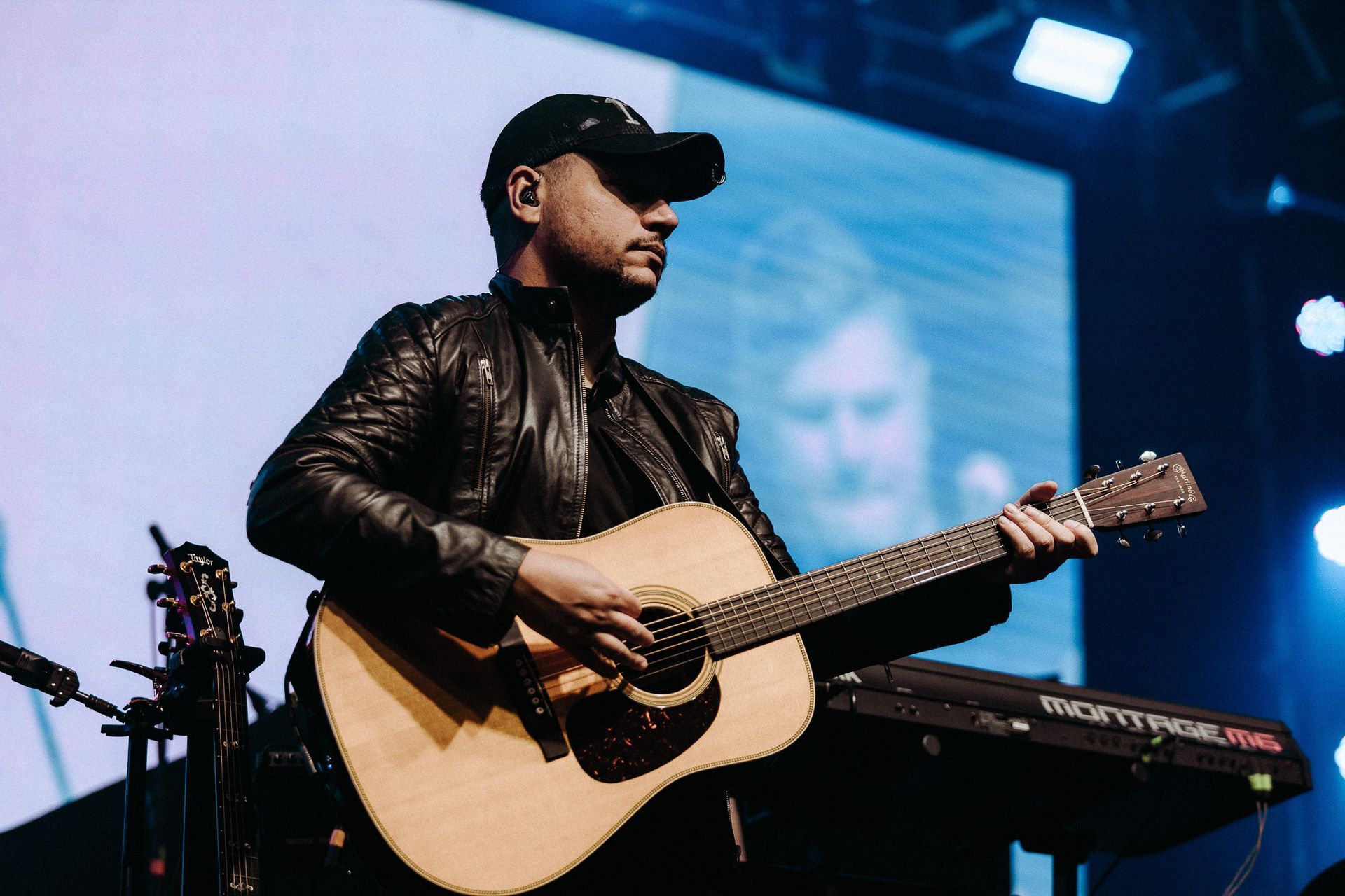 Artista tocando violão no palco, vestindo jaqueta preta e boné de beisebol, com fundo desfocado.