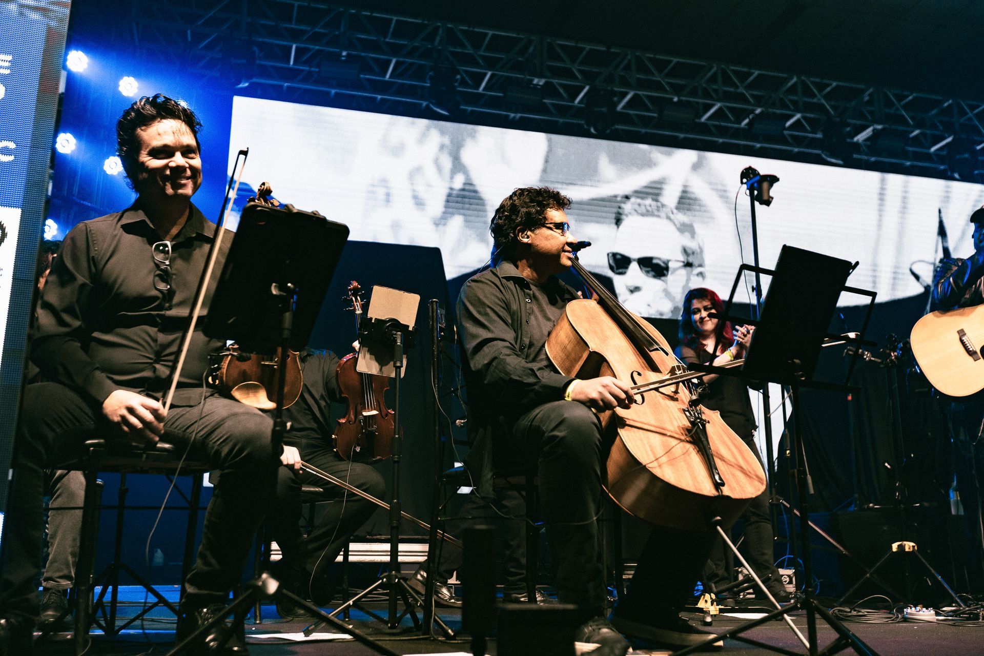 Quarteto de cordas se apresentando no palco com um telão ao fundo. Músicos sentados, alguns tocando, outros sorrindo.
