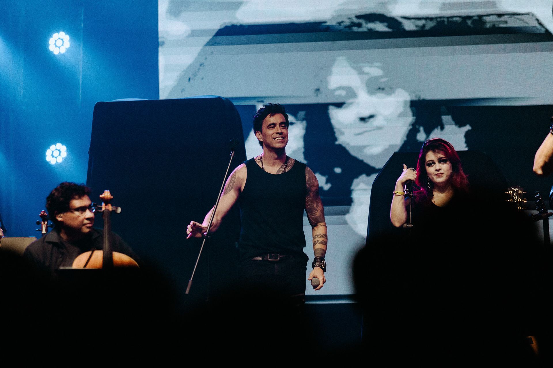 Uma banda se apresenta no palco. Um artista de regata sorri enquanto gesticula. Uma cantora de cabelo rosa