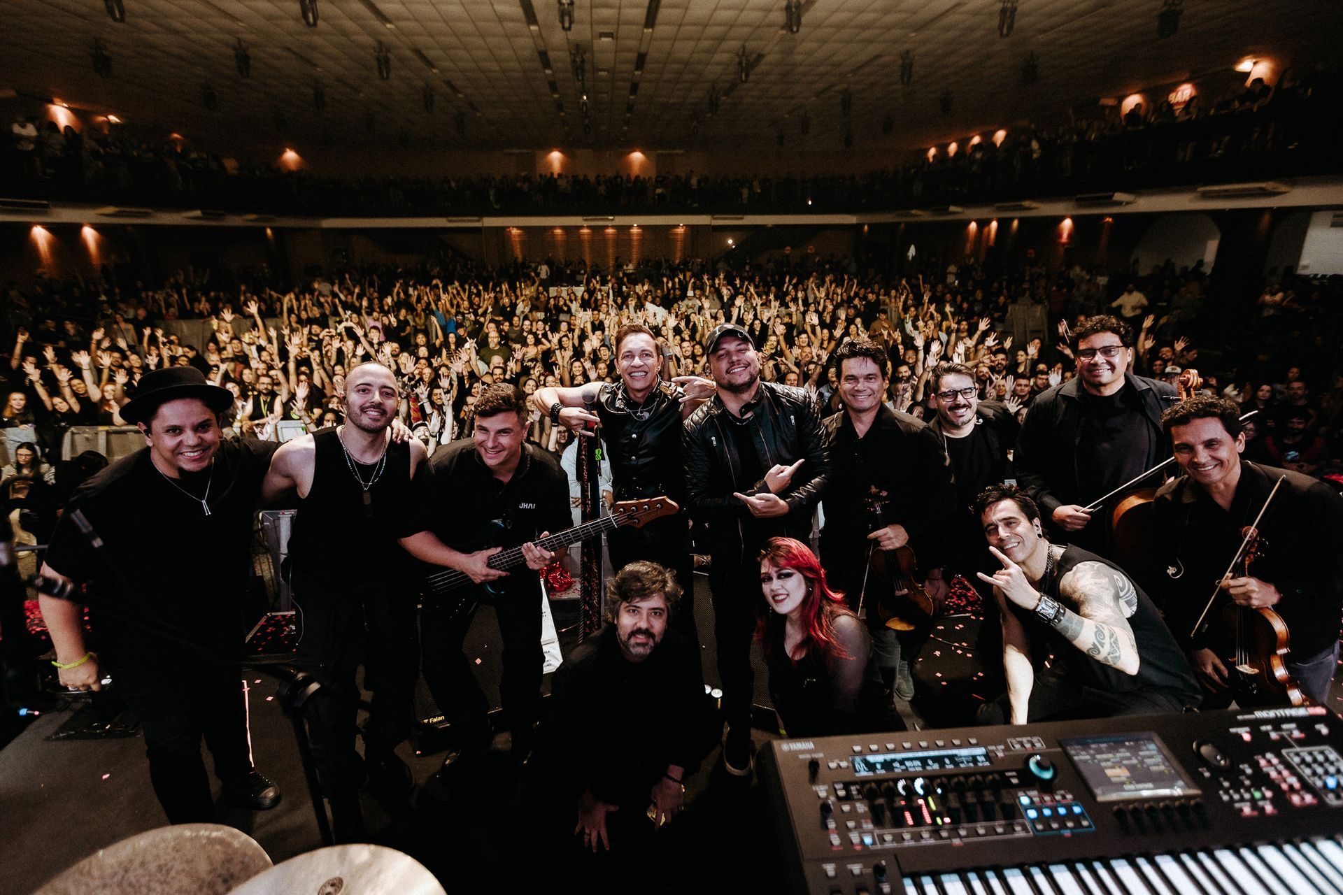 Large band posing onstage with audience behind them and a mixing console in the foreground