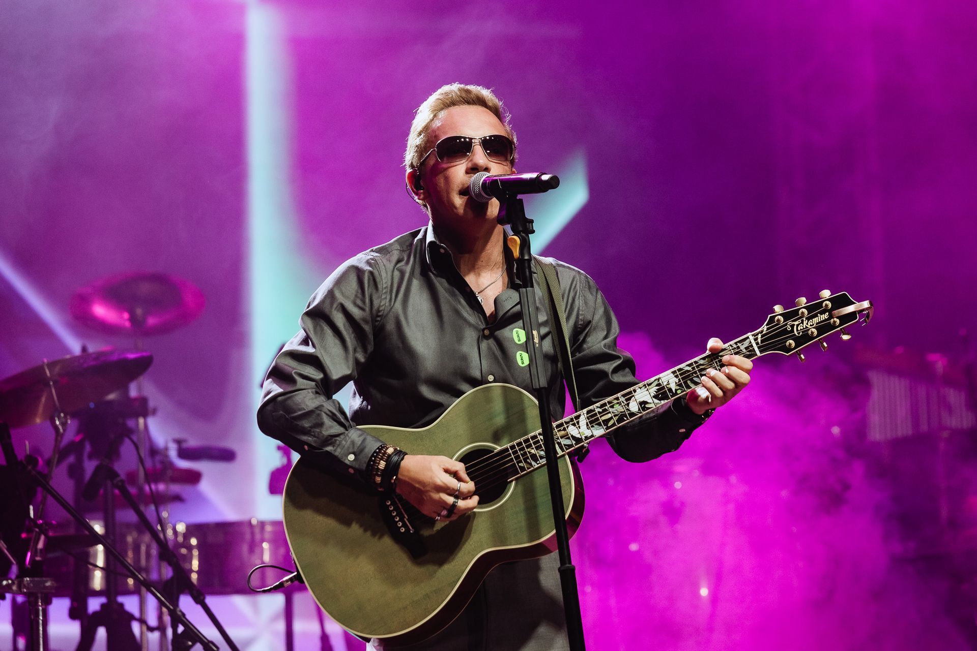 Cantor tocando violão em um palco iluminado de roxo, cantando em um microfone.