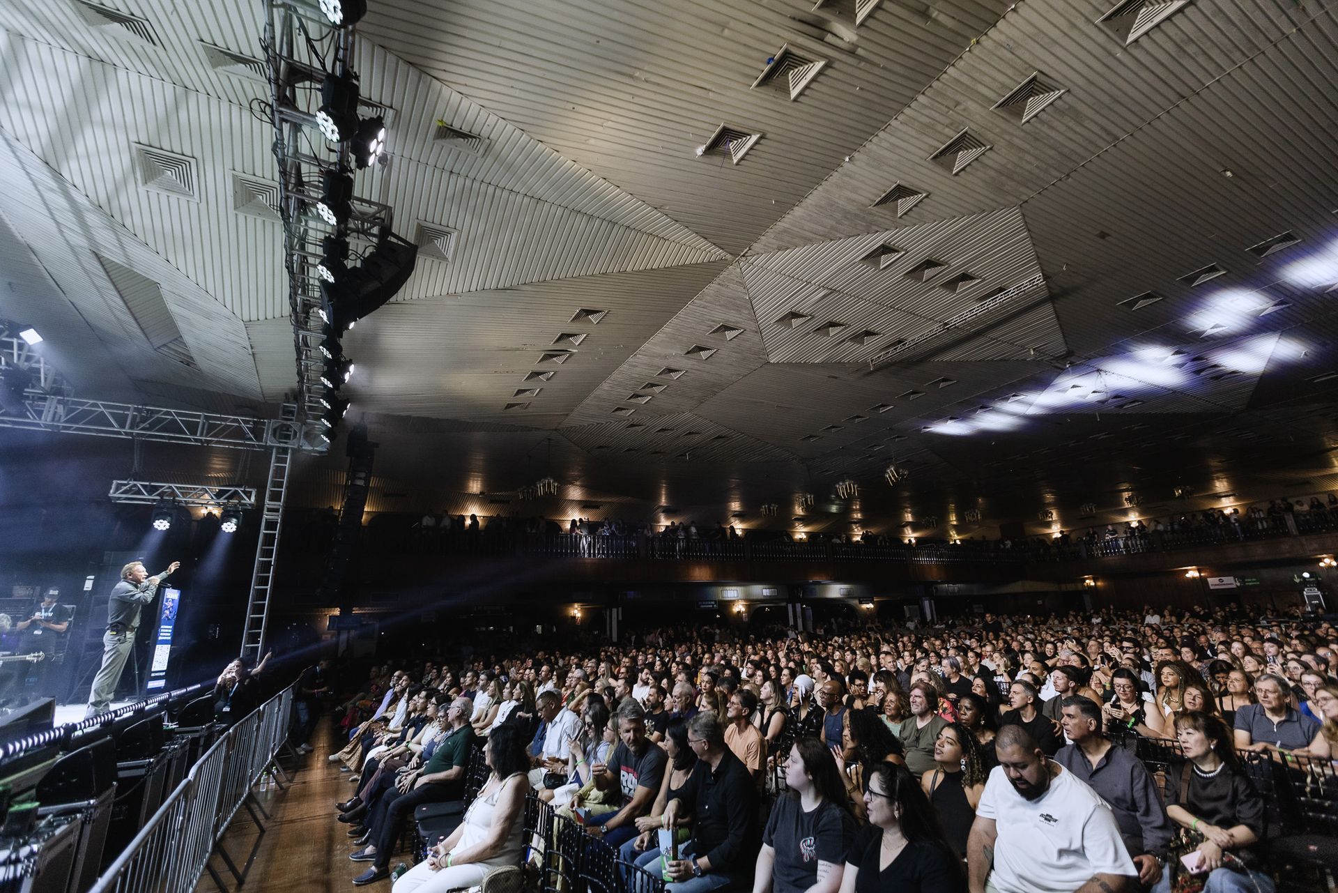 Local de concertos fechado e lotado, com grande público sob um teto texturizado e luzes de palco.