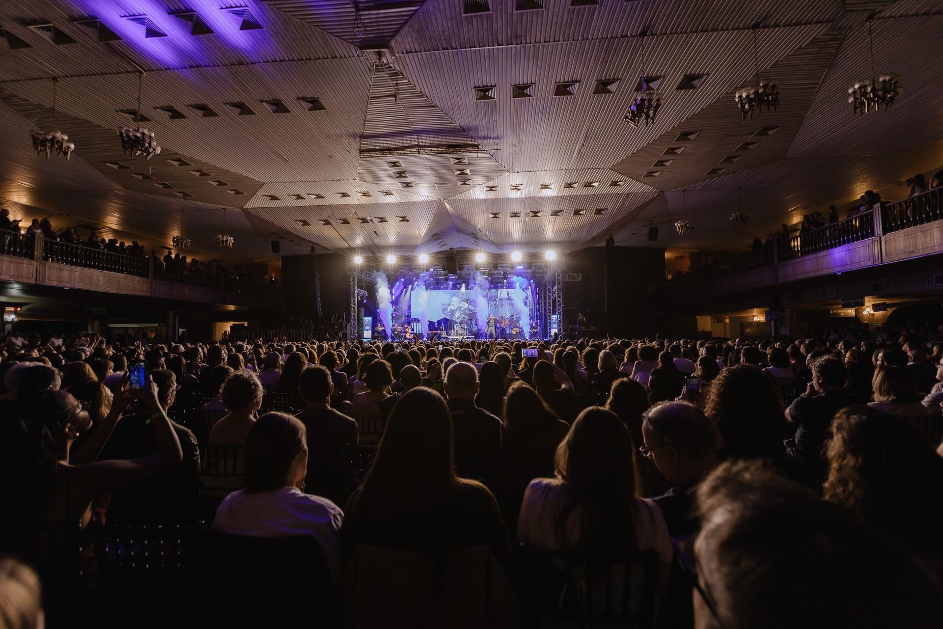 Sala de concertos lotada com o público de frente para um palco intensamente iluminado e luzes roxas no teto.