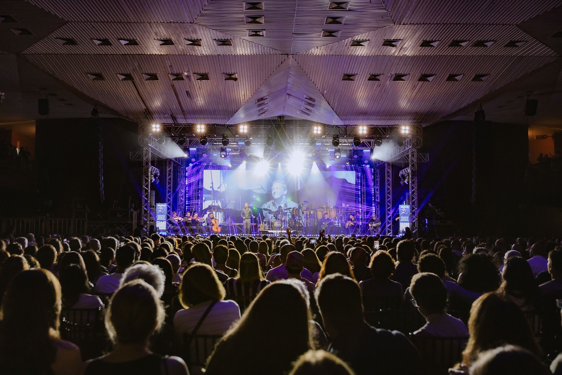 Público assistindo a um show em um palco iluminado em um grande teatro.