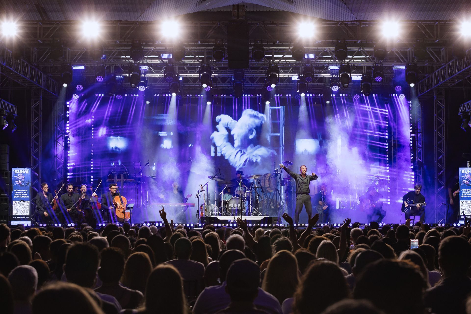 Palco de concerto ao ar livre banhado em luzes roxas, com artistas e público assistindo abaixo.