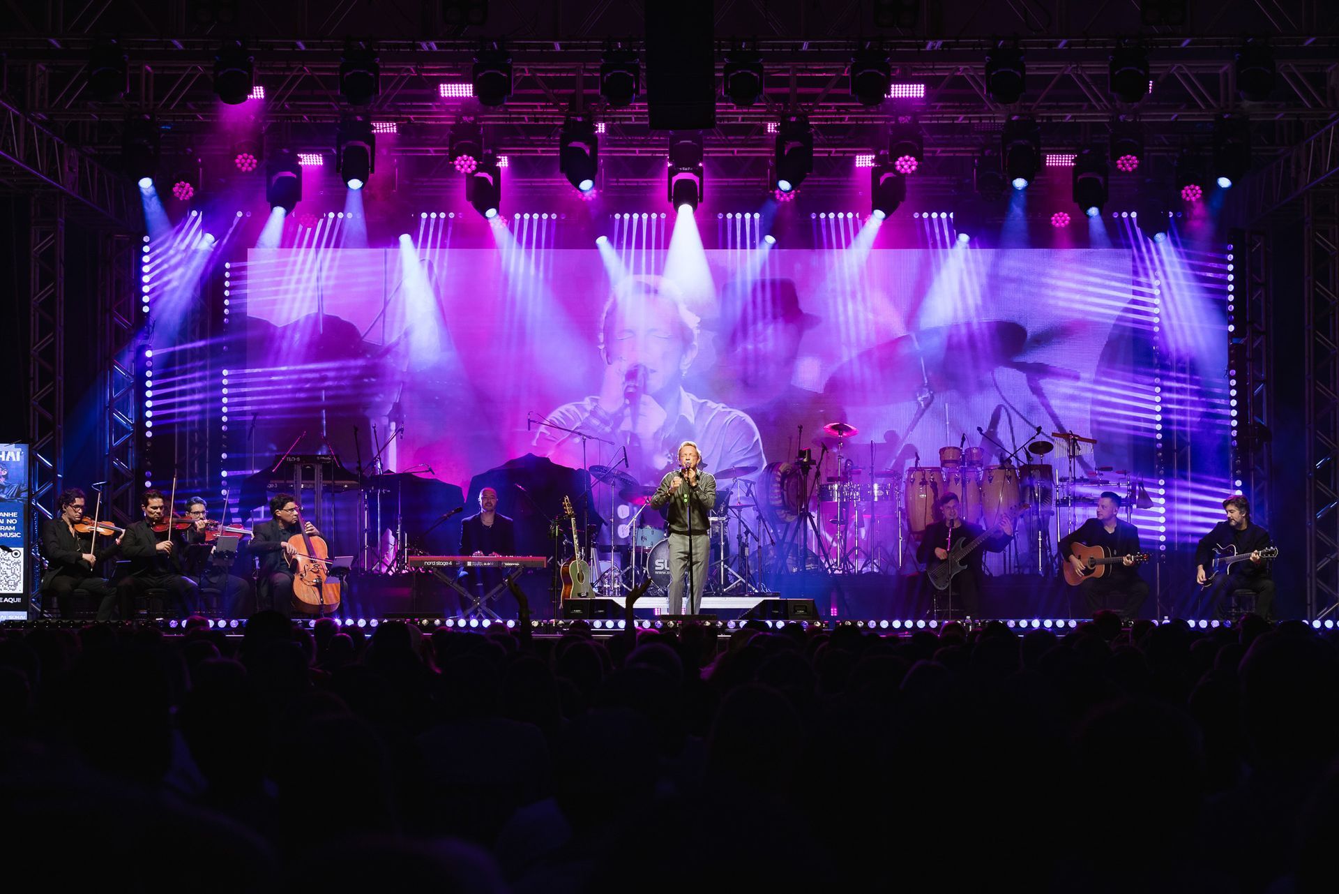 Palco de concerto com luzes roxas, um cantor no centro e uma banda se apresentando diante de um grande telão.